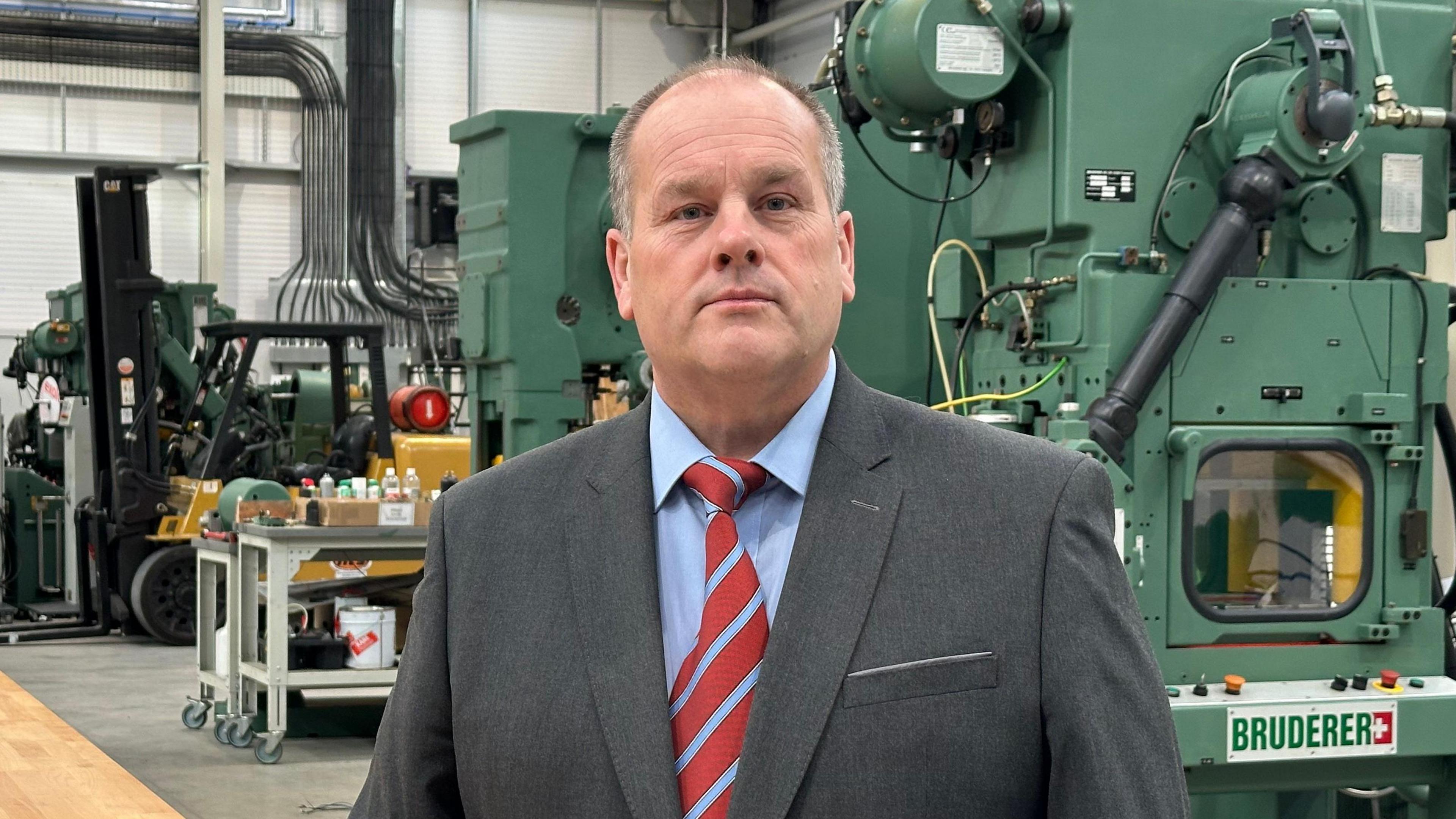 Adrian Haller, wearing a grey suit, blue shirt and a red and blue striped tie, standing in front a big green machine in his factory