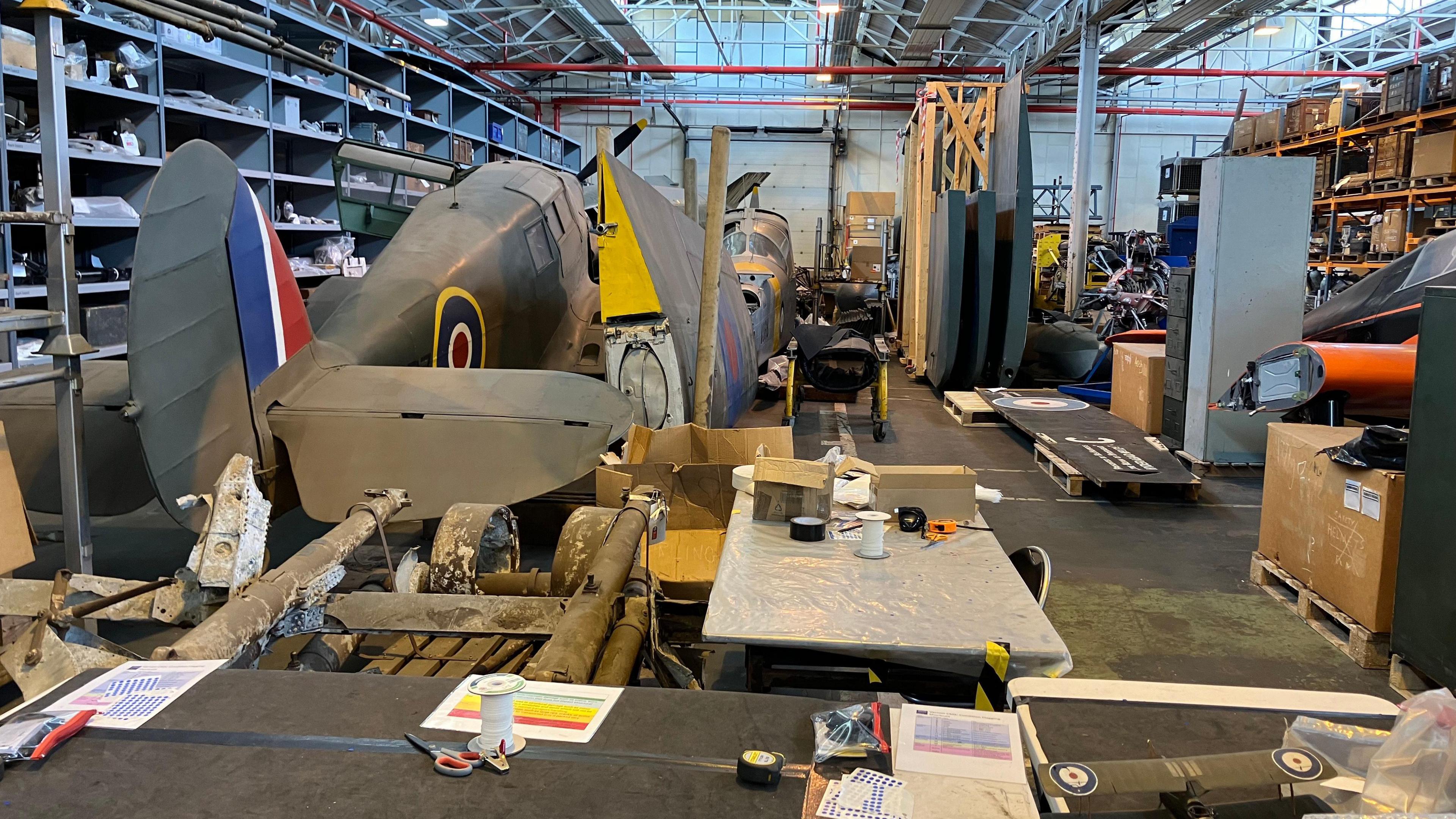 A model of a green RAF plane on the floor of a warehouse, with other models and equipment around it. Shelves containing boxes and other items are at the side of the warehouse.