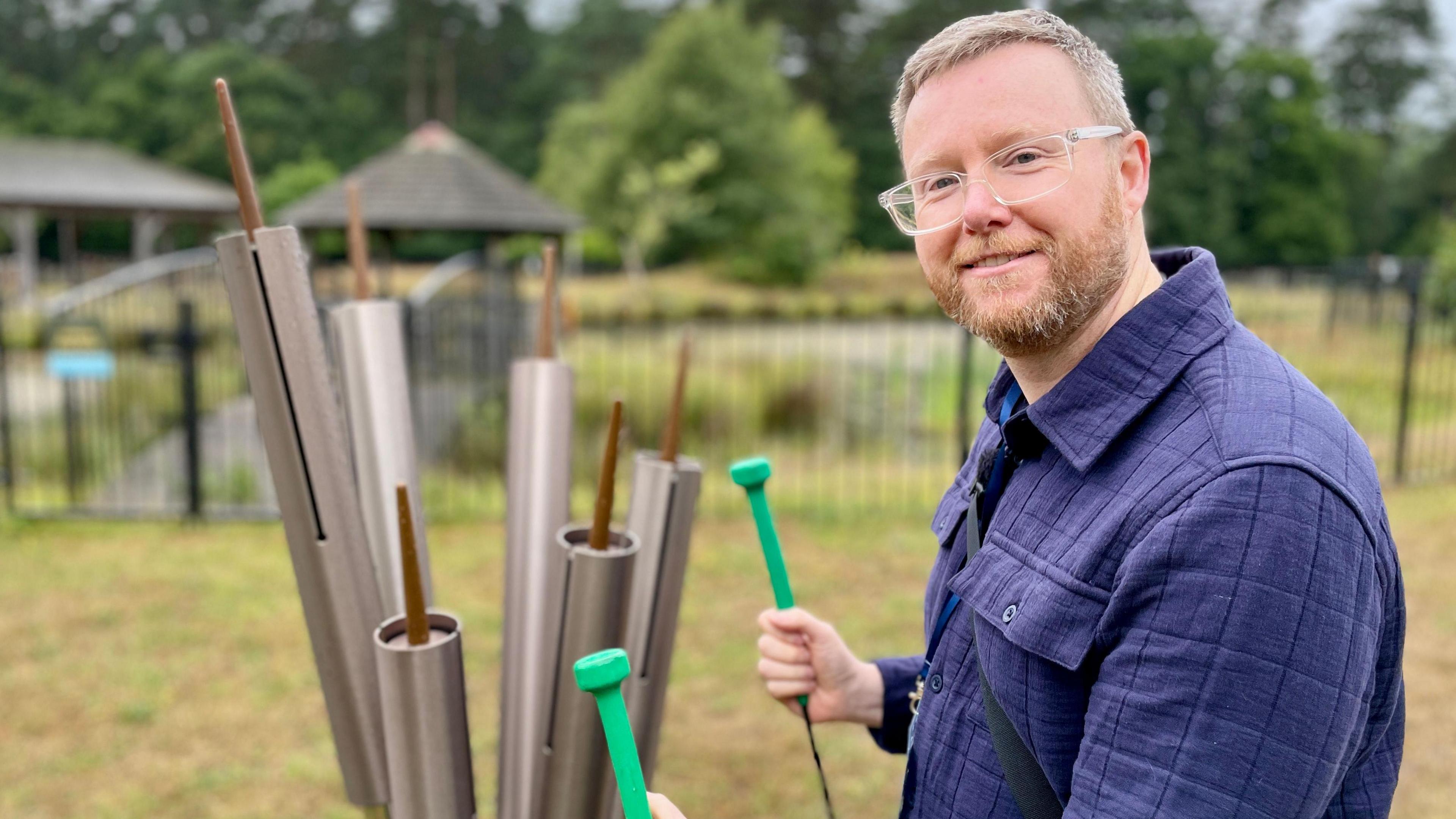 Stuart Wood stands next to a set of tubular bells with a pond behind him while holding two chime hammers.