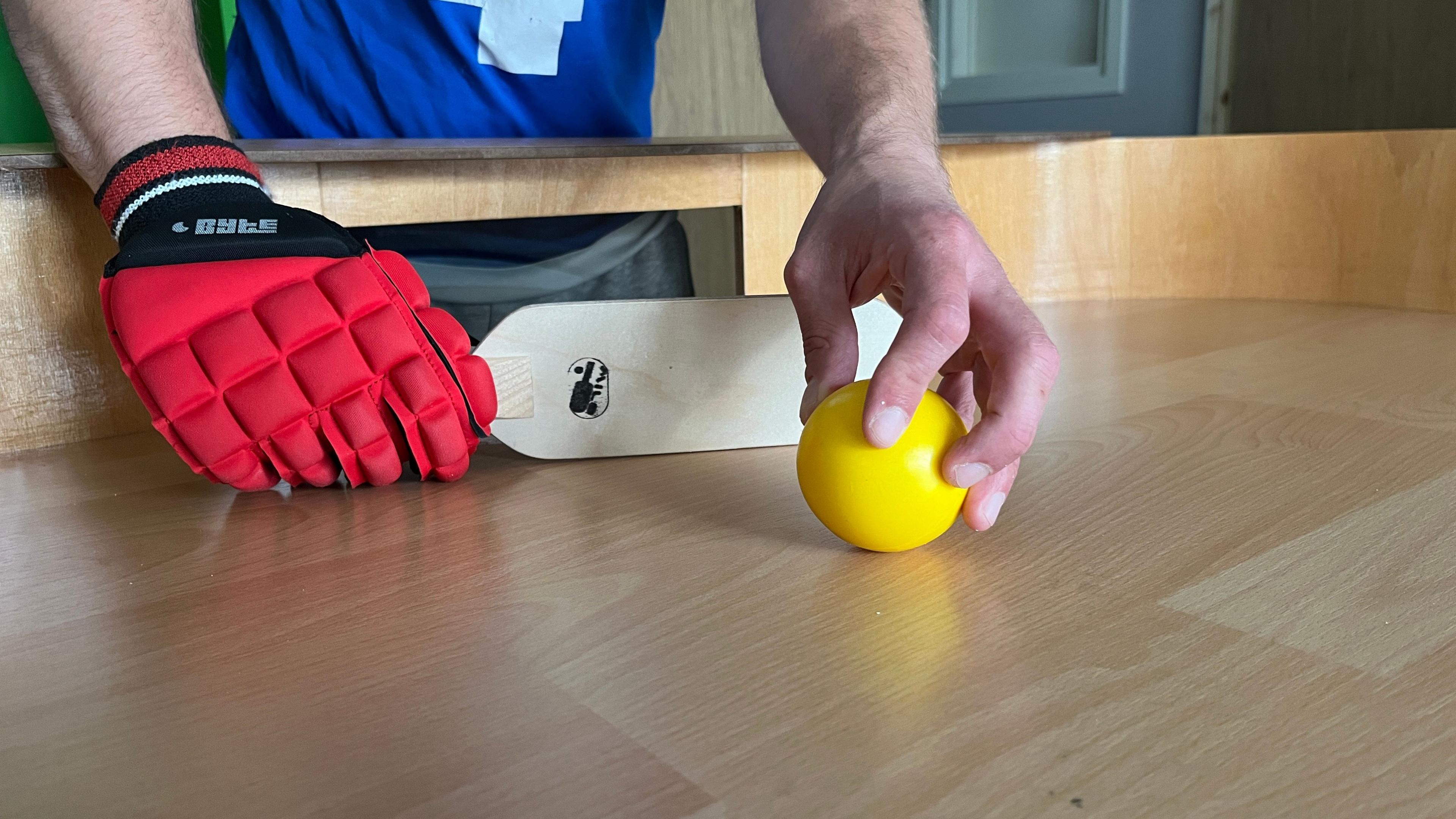 A close up of Mr Wilson's hands on a Showdown table. In one hand, he holds the bright yellow ball and the in other he holds a small, wooden flat bat. On the hand holding the bat he is wearing a padded red glove.