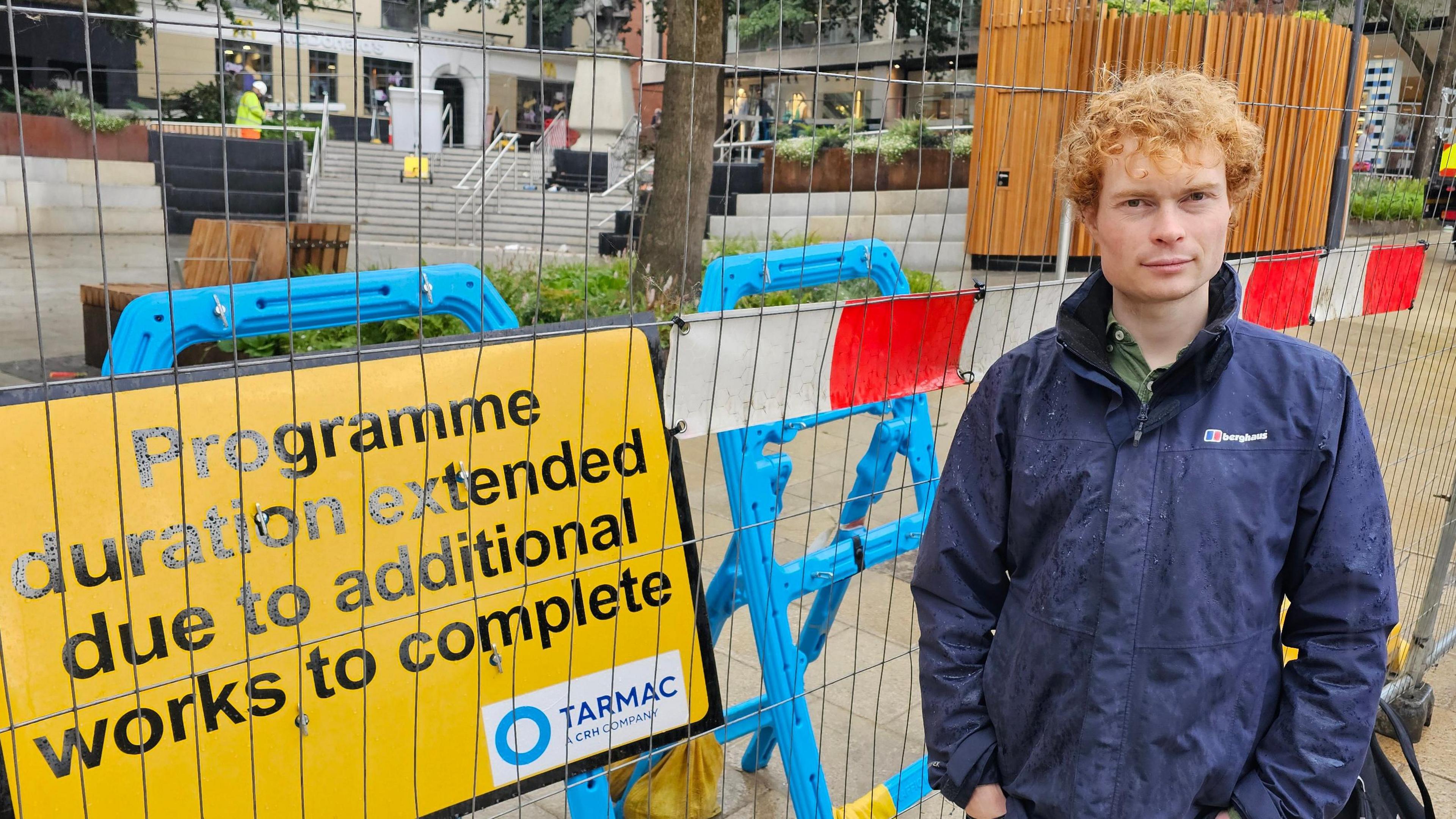 Jamie Osborn standing next to fencing surrounding Hay Hill, along with a sign which says 'Programme duration extended due to additional works to complete'.