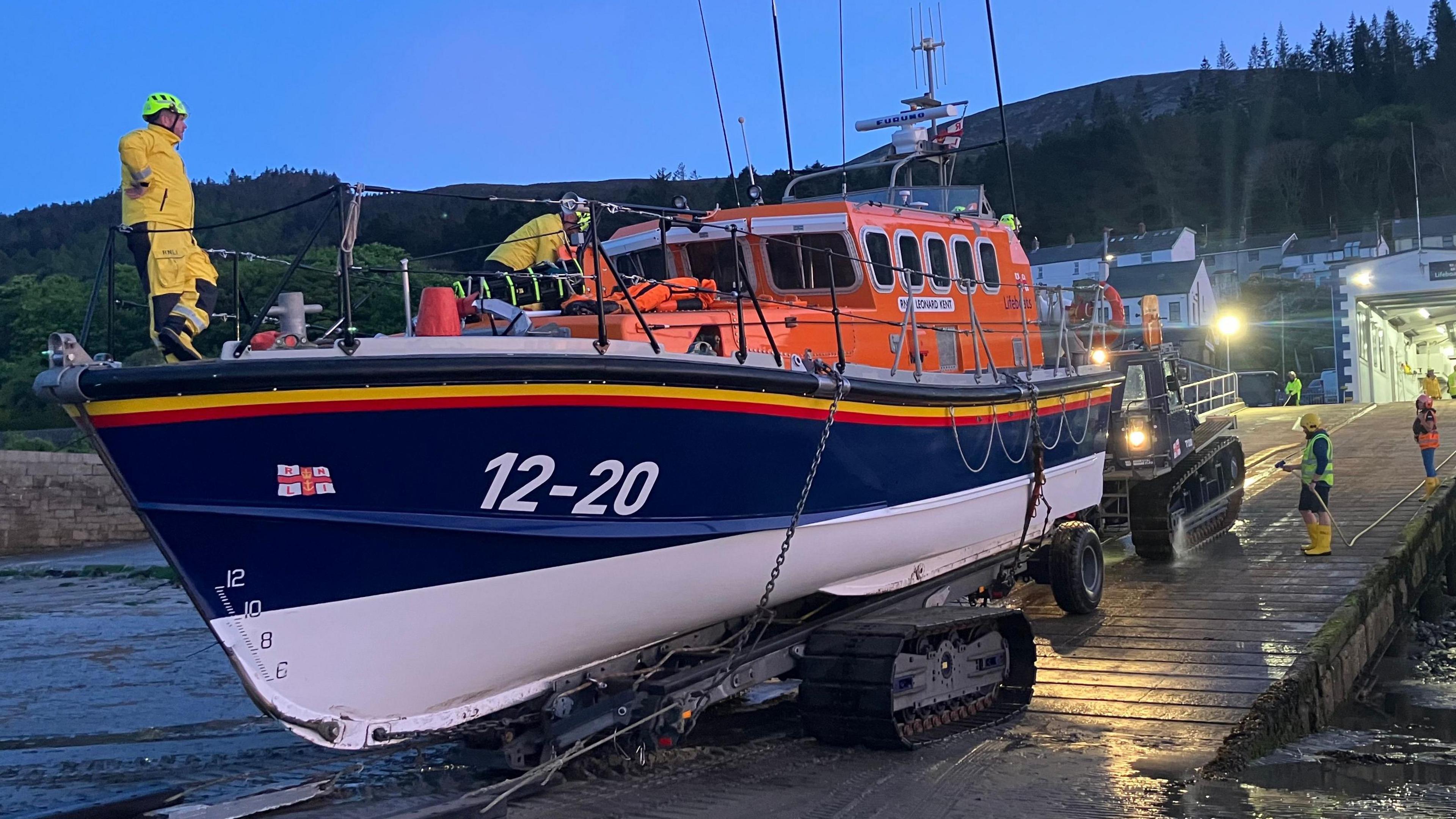 An orange, blue and white boat that is docked in the evening on a ramp with men in yellow standing on the boat and deck and fixing maintaining it.