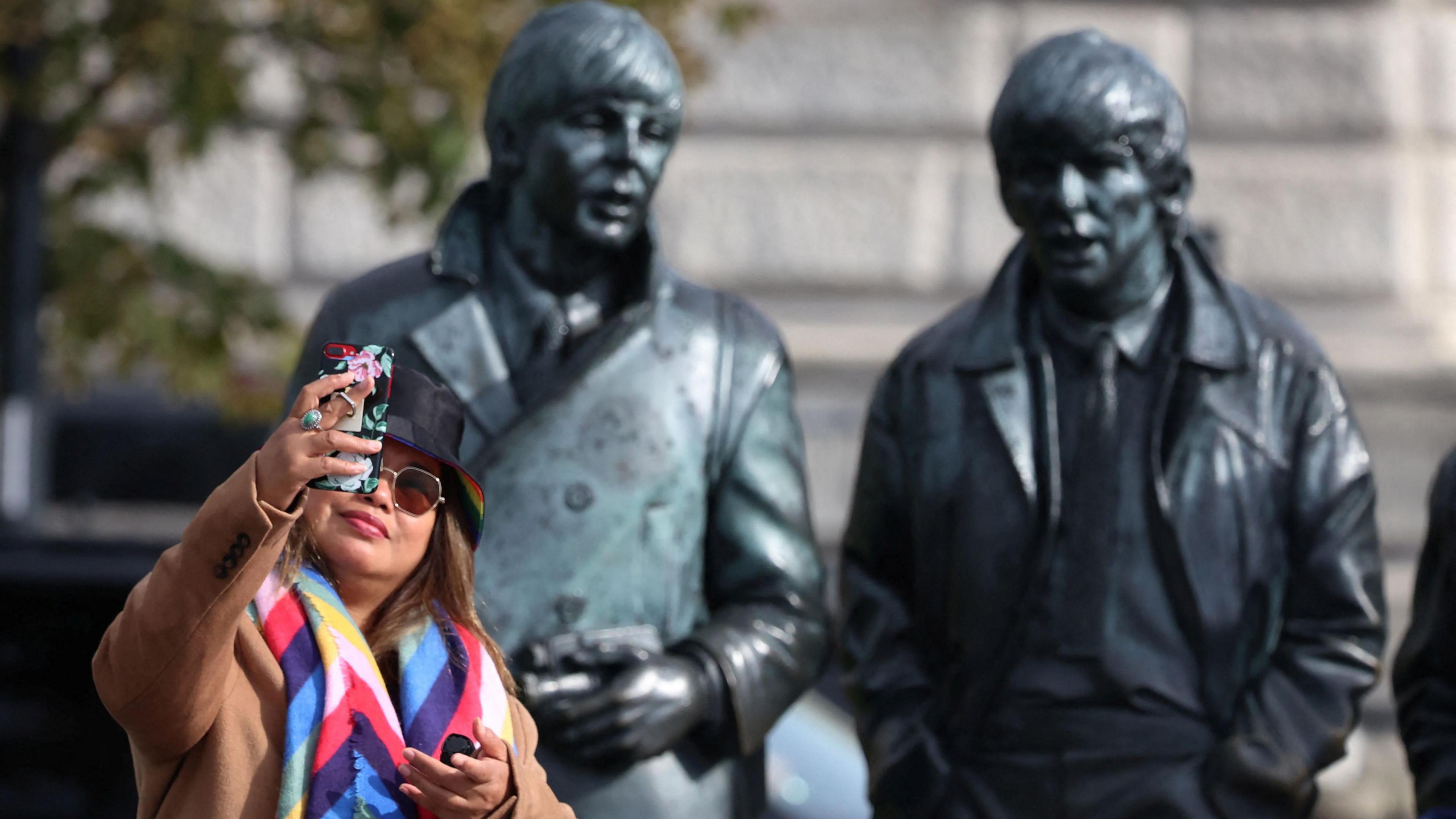 Woman in hat, shades and bright multi-coloured scarf takes a selfie in front of the Beatles sculpture, showing Paul McCartney and George Harrison in the backdrop