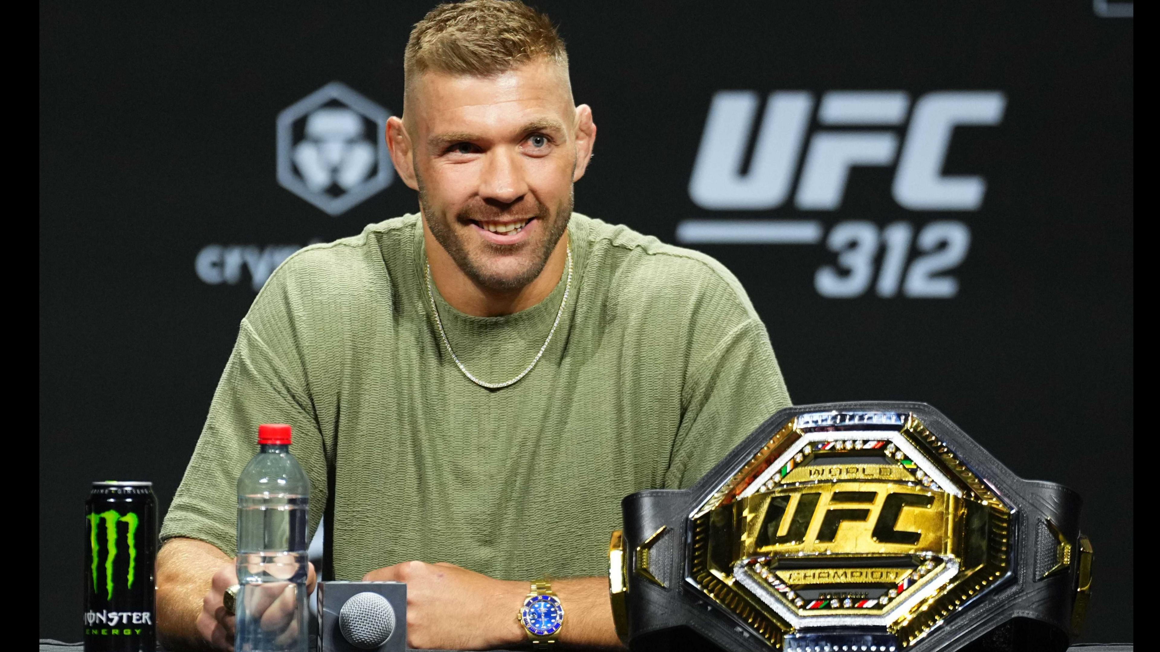 Dricus du Plessis, wearing a green t-shirt with a metal chain, poses at a desk with a microphone and his UFC middleweight belt in front of him