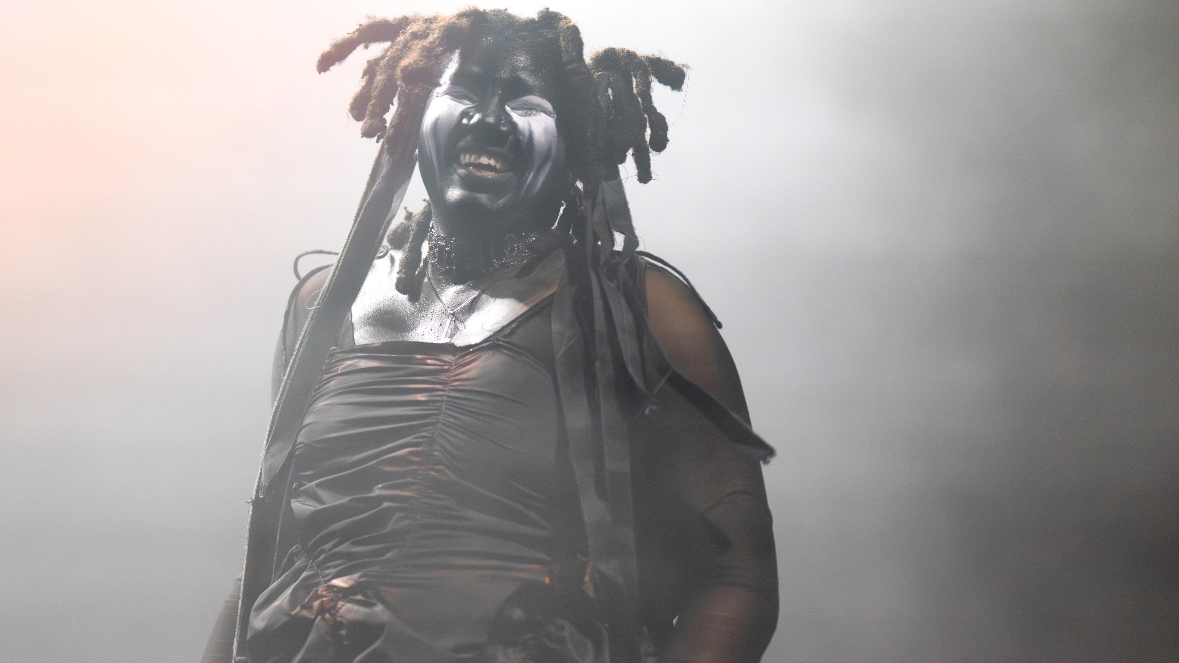 Artist Backxwash has thick dreadlocks with black ribbons attached to some, black and white face paint, and a ruffled black top