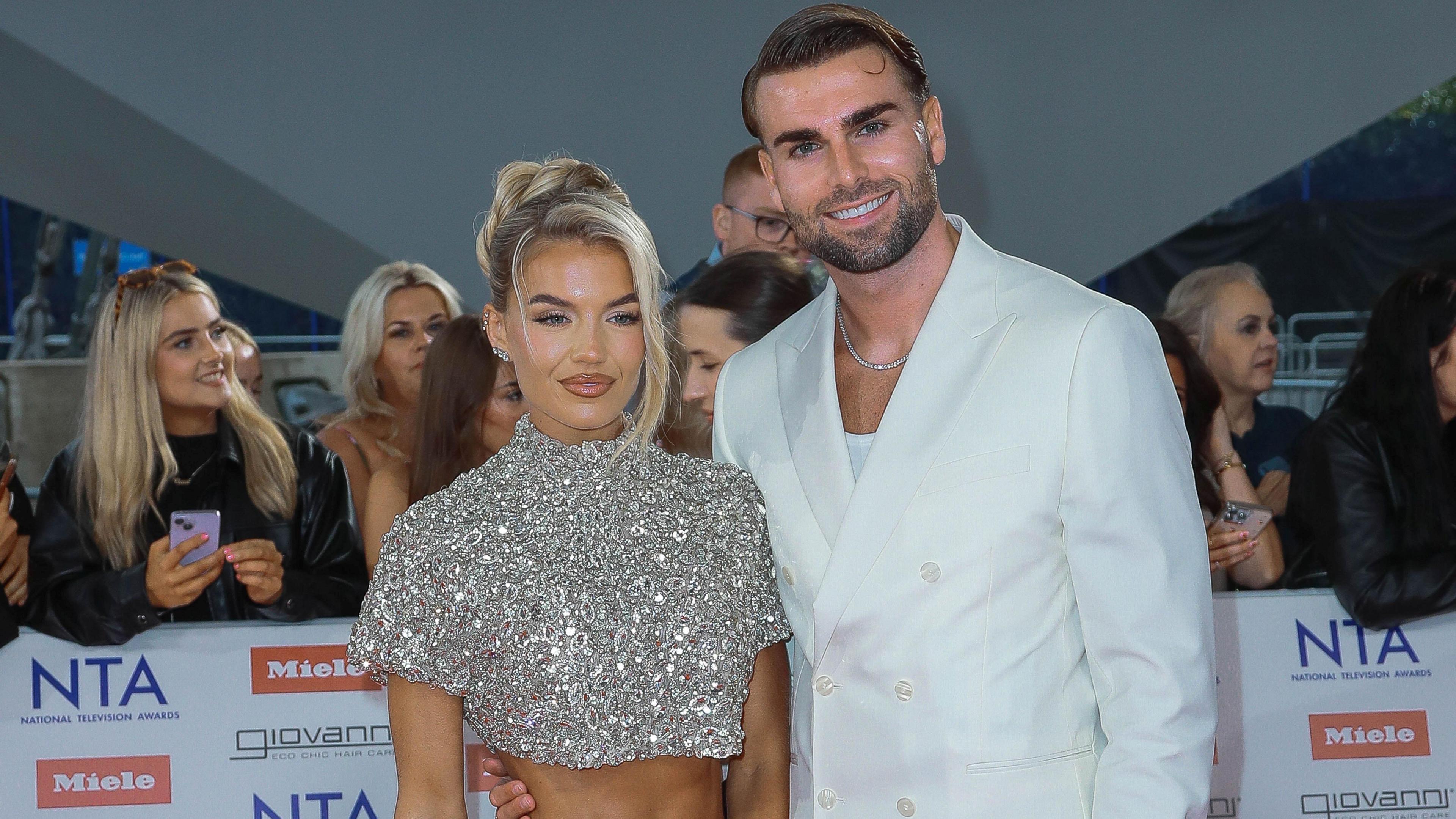 Molly Smith and Tom Clare post together on a red carpet. Molly is wearing a sparkly cropped top and Tom is wearing a white suit over a white top.  