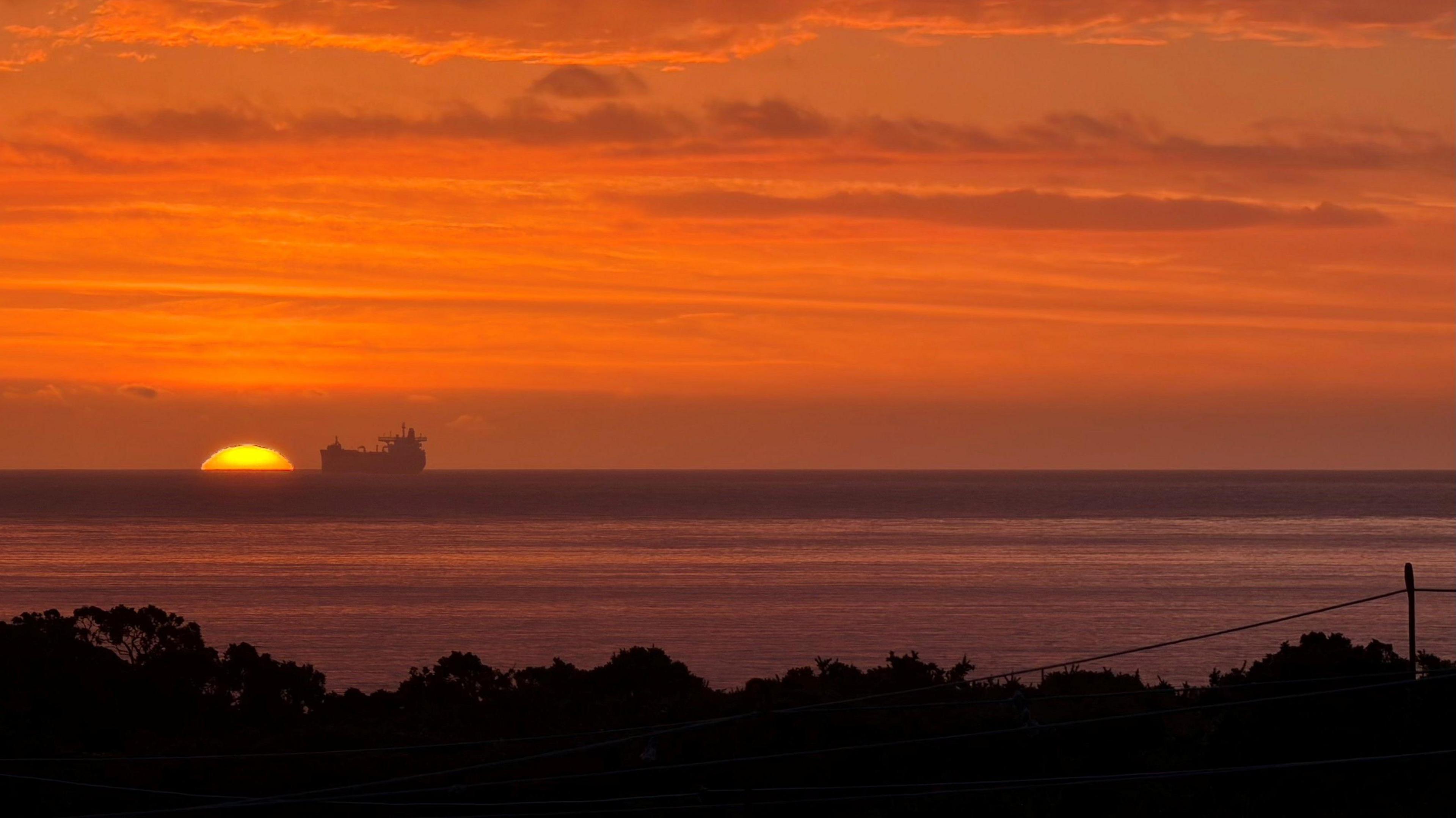 An image of an orange sunrise sky, with the sun appearing over the North Sea horizon, next to an offshore vessel in the distance.