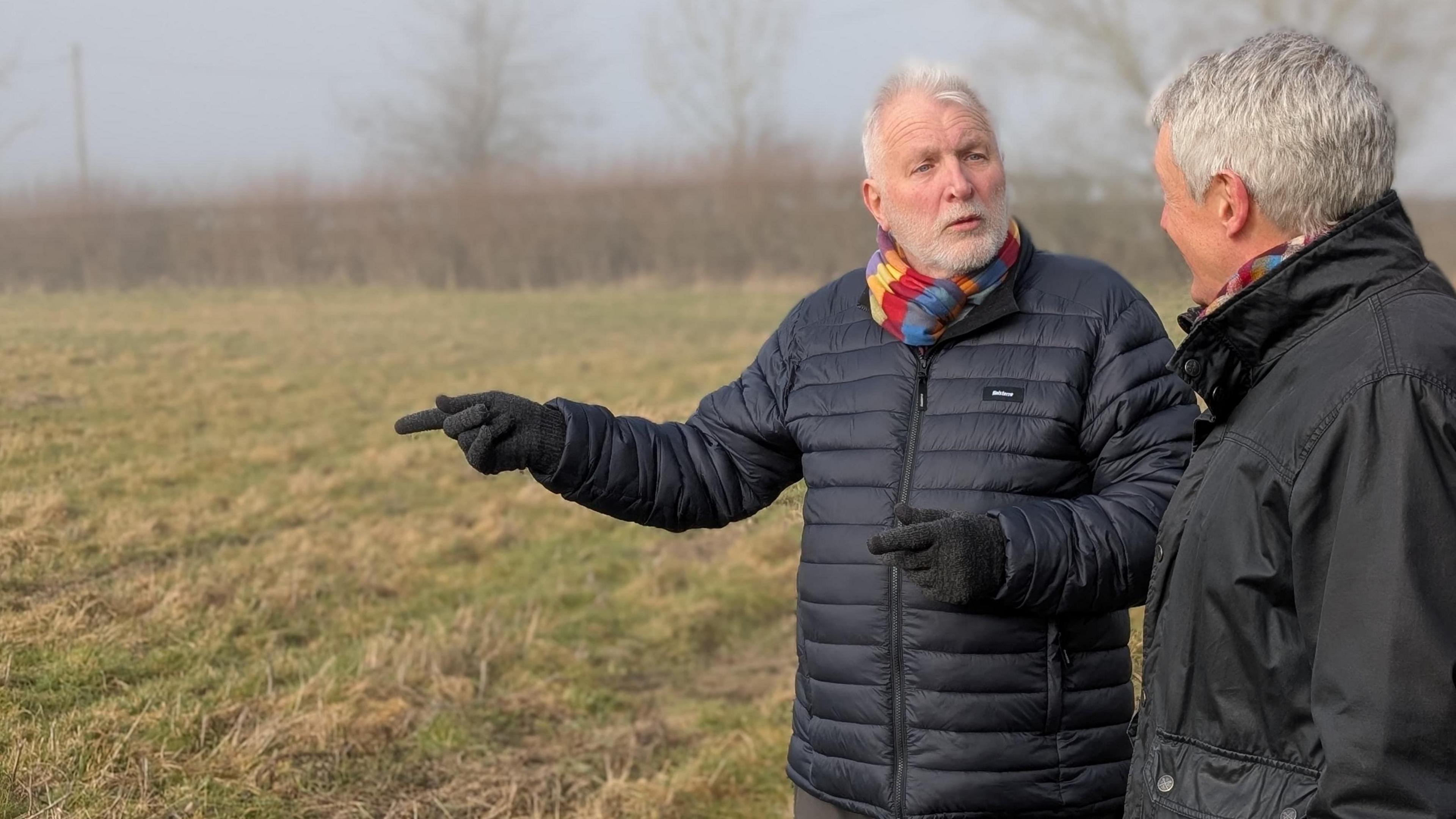 David Still and Nick Hayward, both in their 60s, are wearing dark jackets and colourful snoods. They're standing talking and gesturing in a field and there is a misty grass verge behind them. 