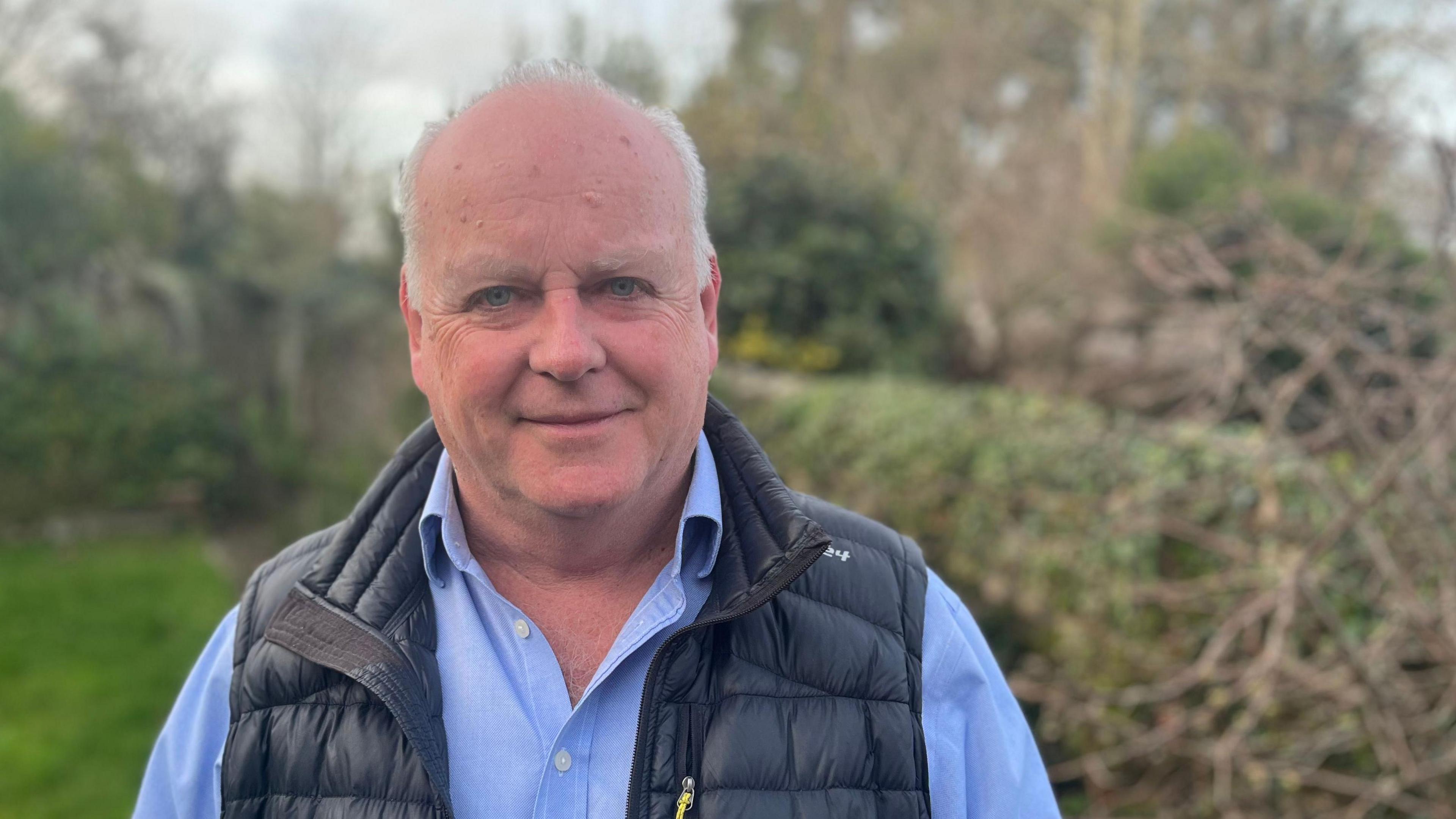 Paul Arnott, deputy leader of Devon County Council, wearing a light blue shirt and a navy blue gilet and standing in countryside with a blurred out autumnal hedge behind him.