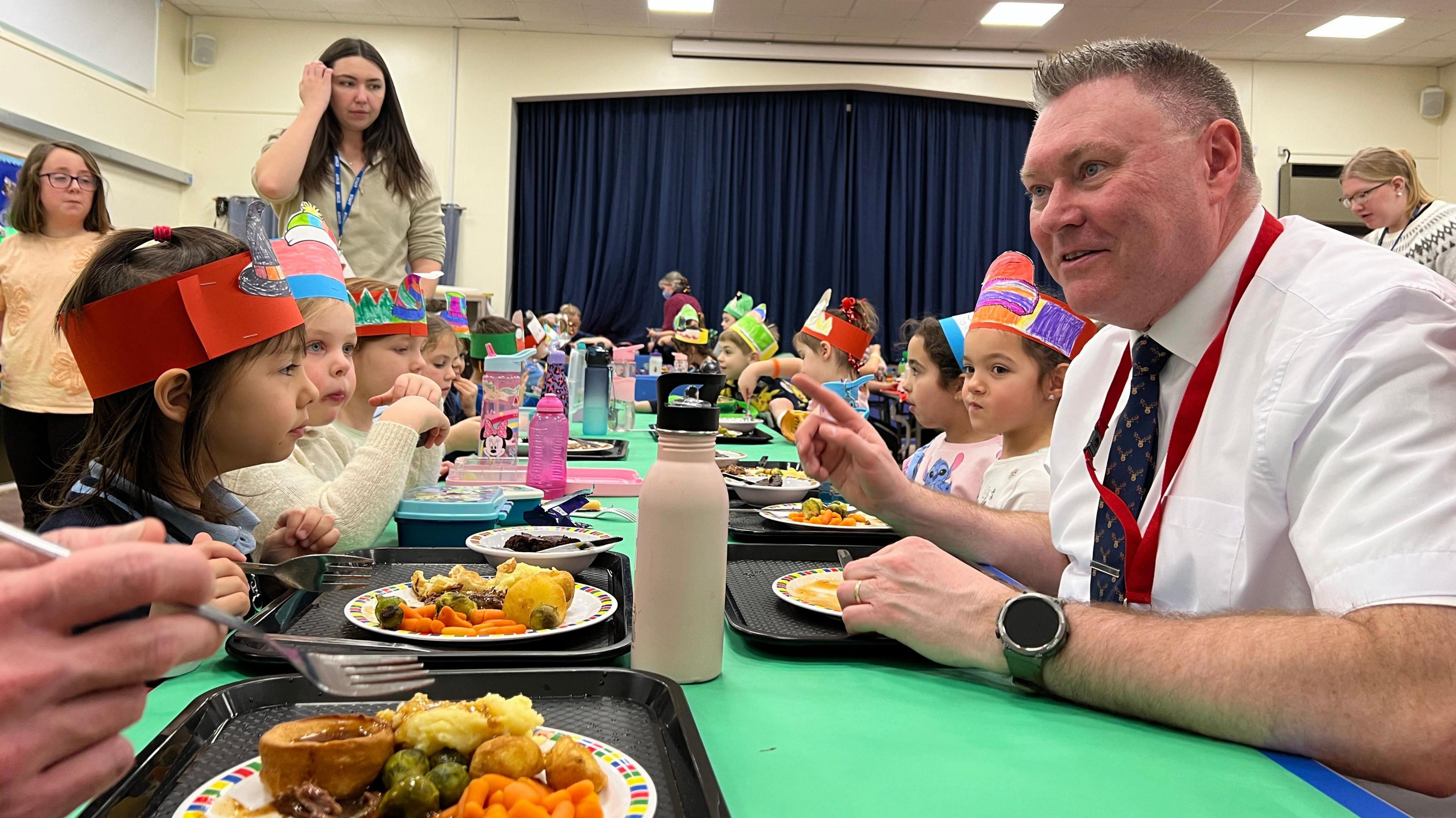 Rob Ward sits on a children's table with pupils and eats his lunch. He's wearing a white shirt with a dark blue tie and his sleeves are rolled up. He has short grey hair which has been spiked up. The children around him are wearing cardboard decorated hats on their heads and their trays of food are in front of them.