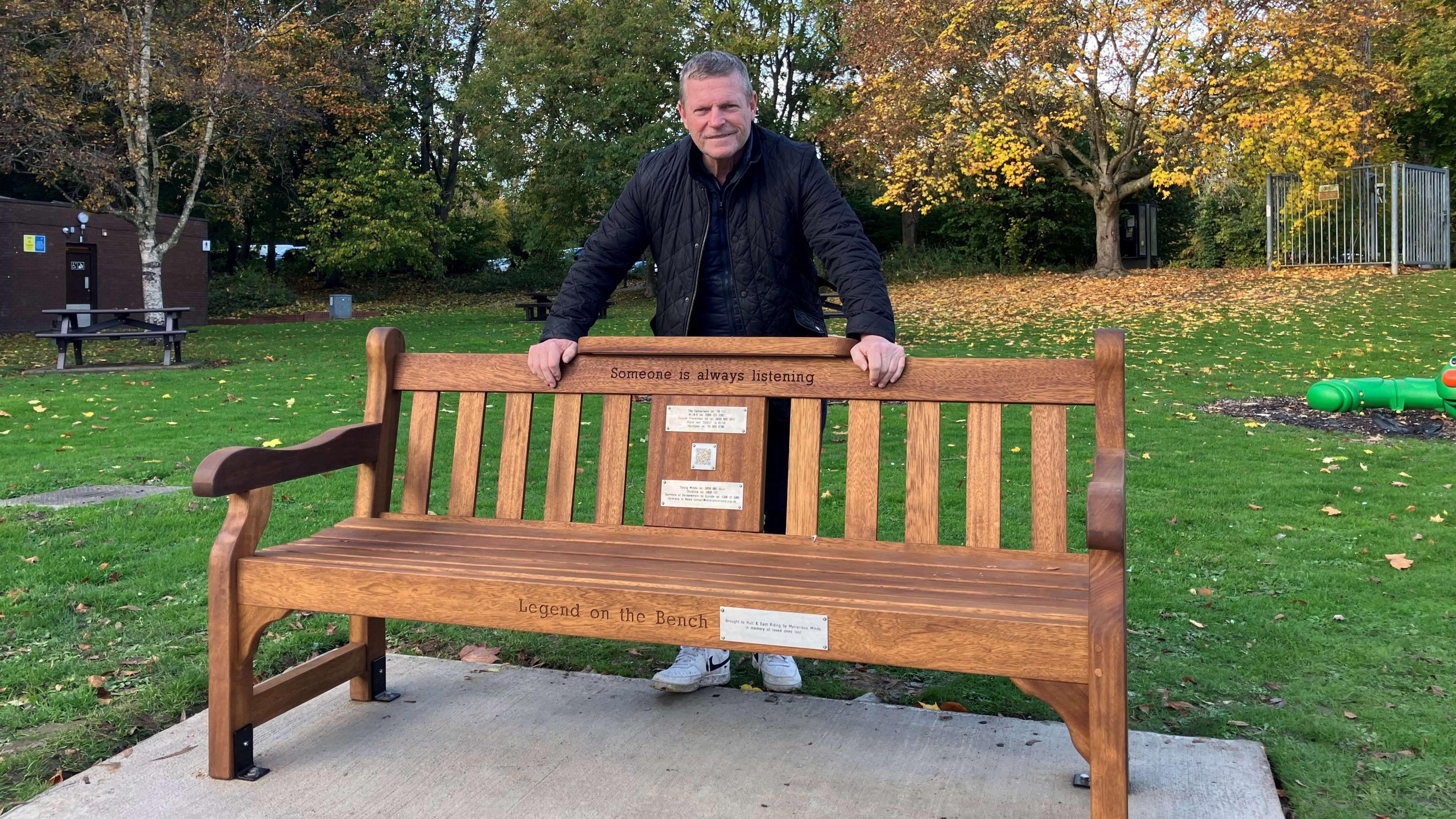 A man with a black jacket and white trainers leaning on a wooden bench in a grass area.