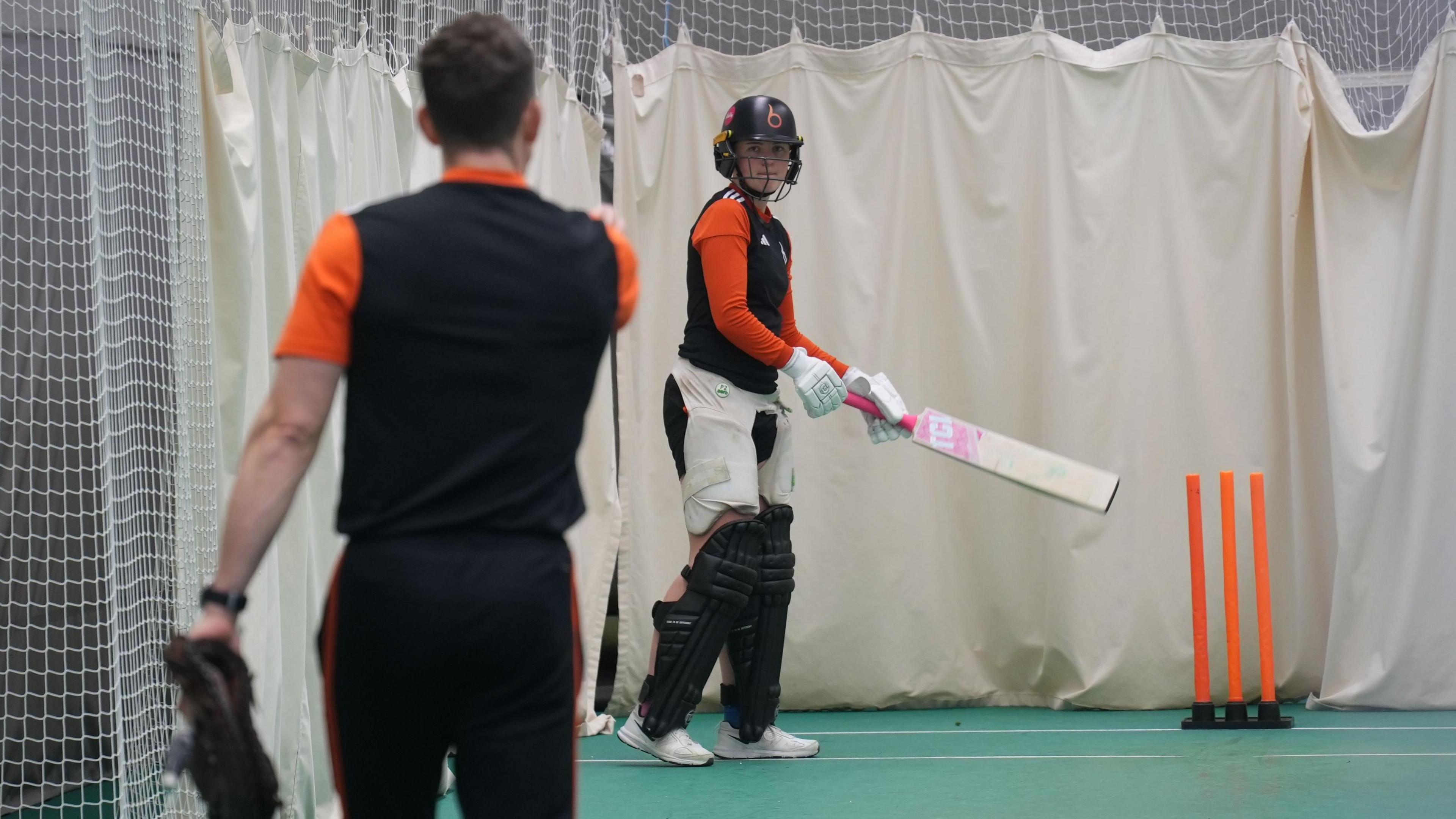 Grace Ballinger stands with bat in hand listening to a coach give her feedback in training. She has full batting kit on and is in indoor nets at Trent Bridge.