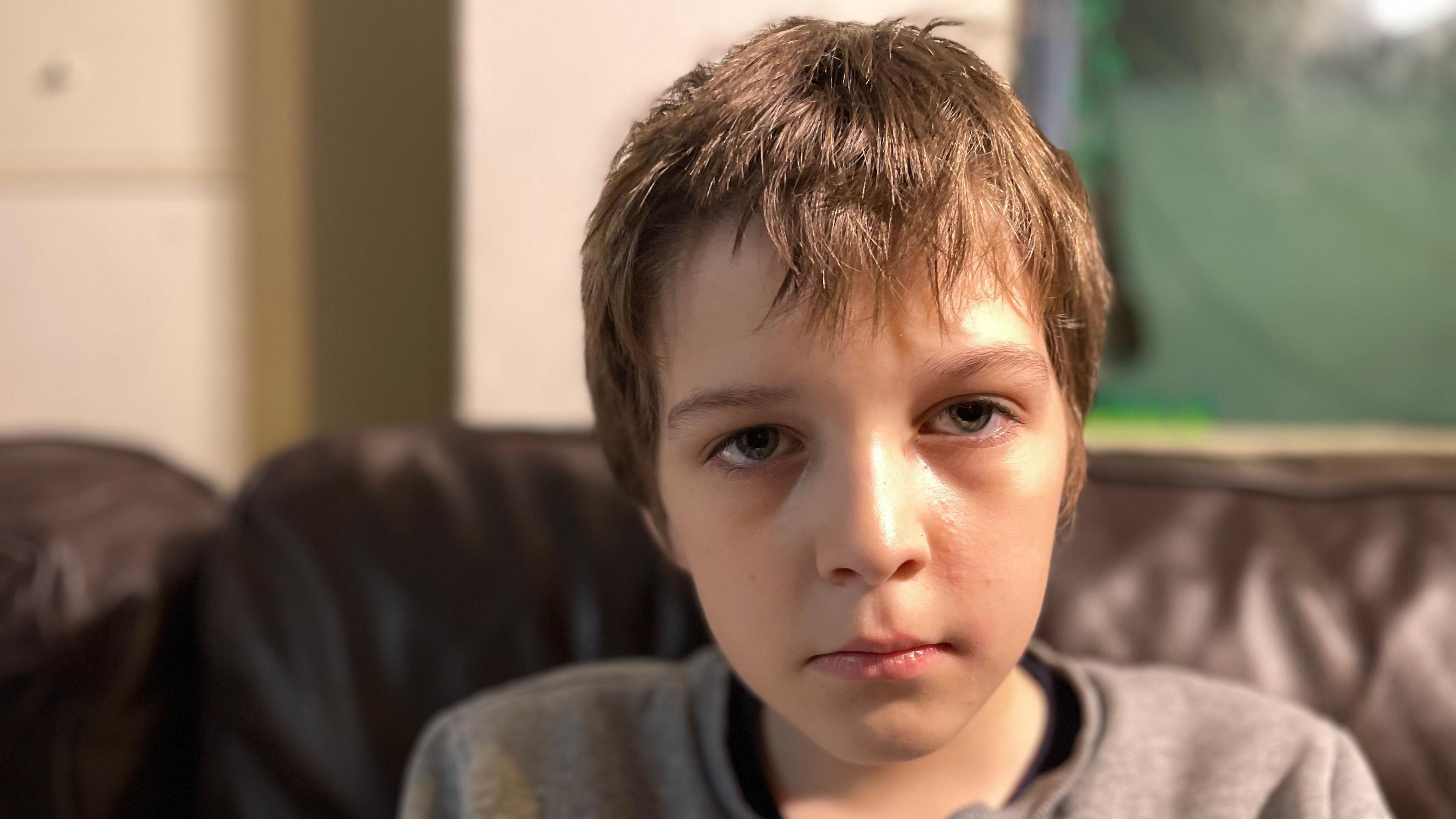 Glum-looking 12-year-old boy in a grey jumper sitting on a brown leather sofa