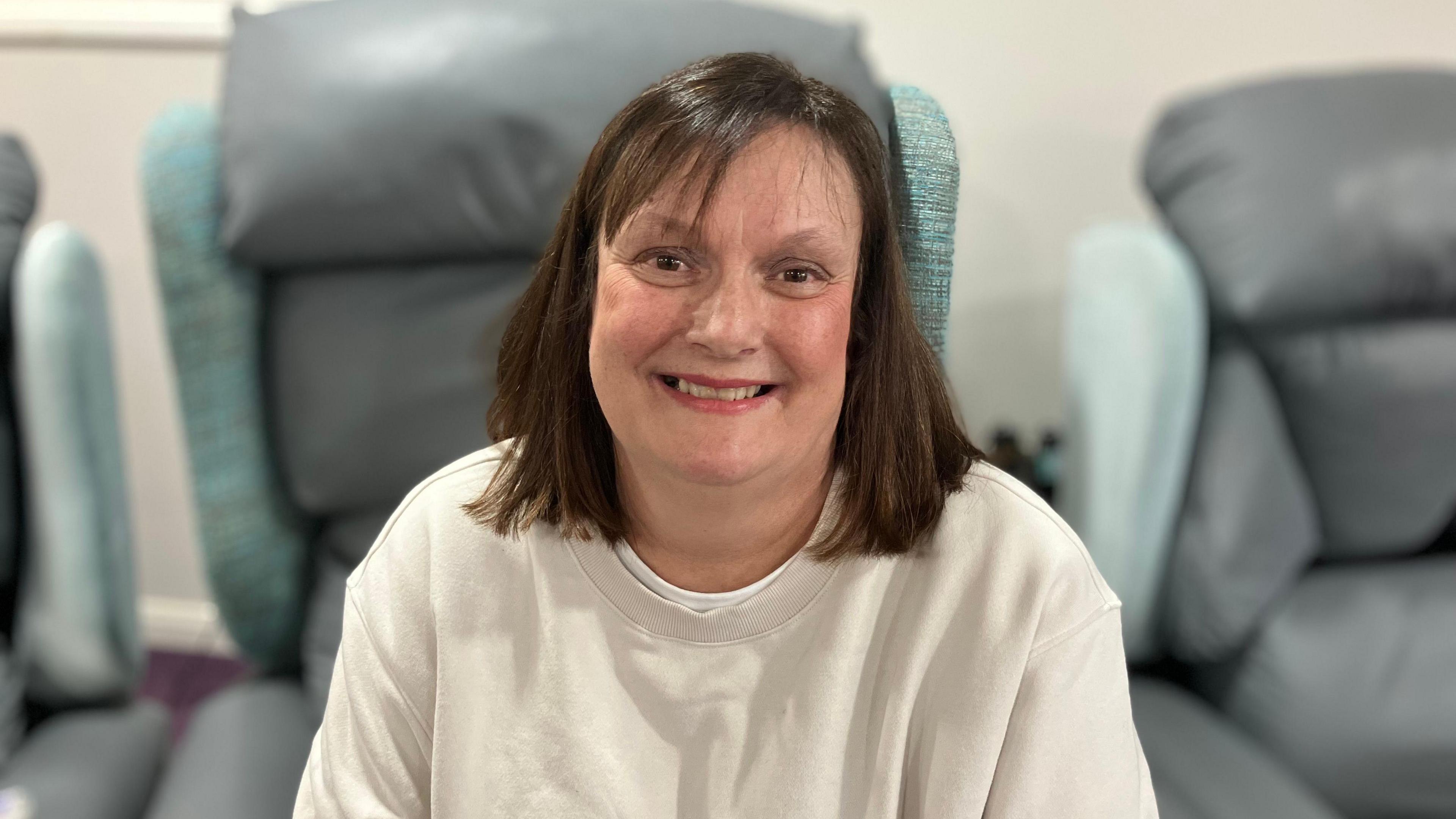 A woman looking towards the camera and smiling. She is sat on a large, grey leather chair. She is wearing a beige sweatshirt. 