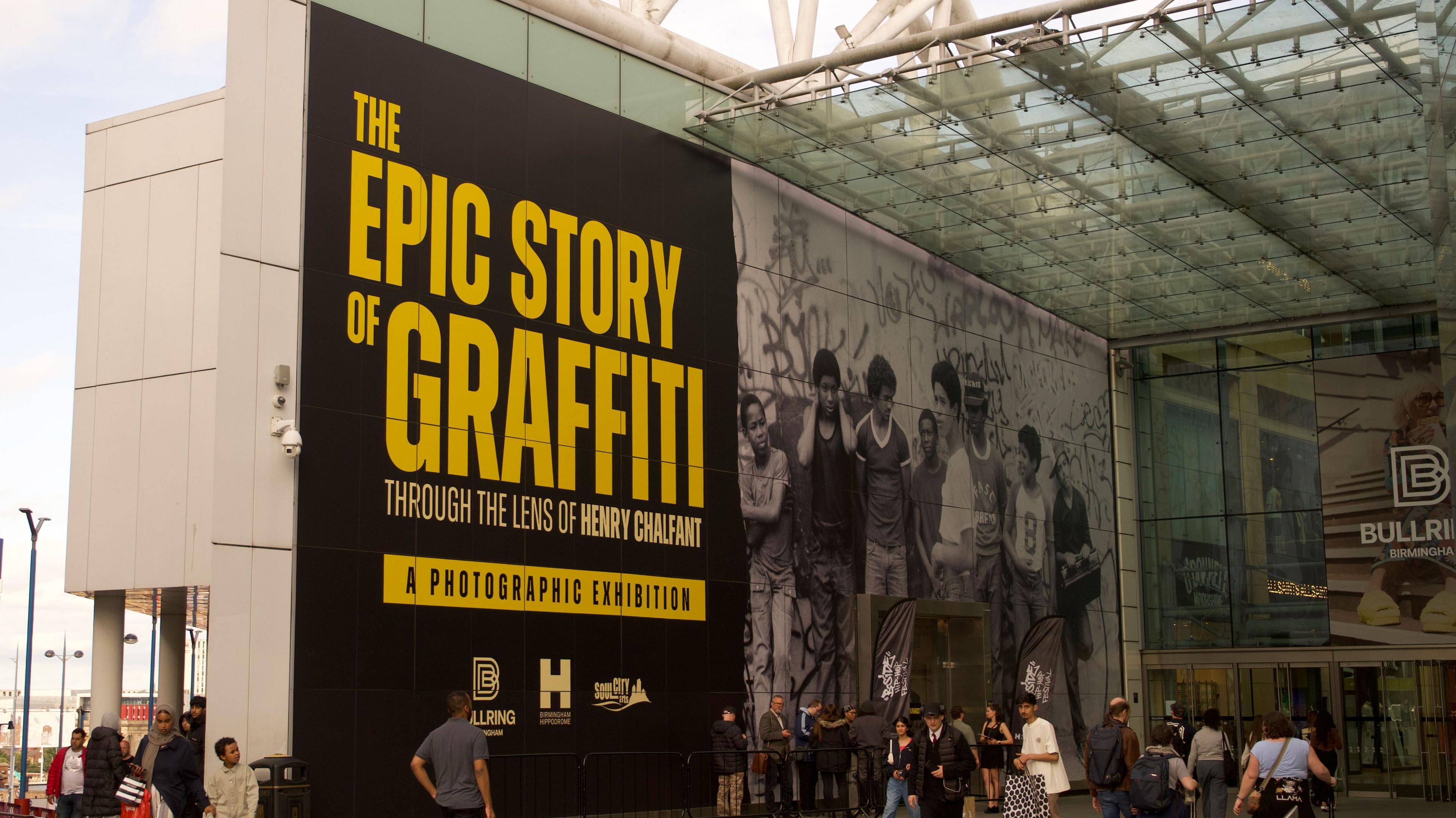 A giant image with the words 'The Epic Story of Graffiti: Through the lens of Henry Chalfant' in yellow on the side of an entrance to a modern shopping centre.