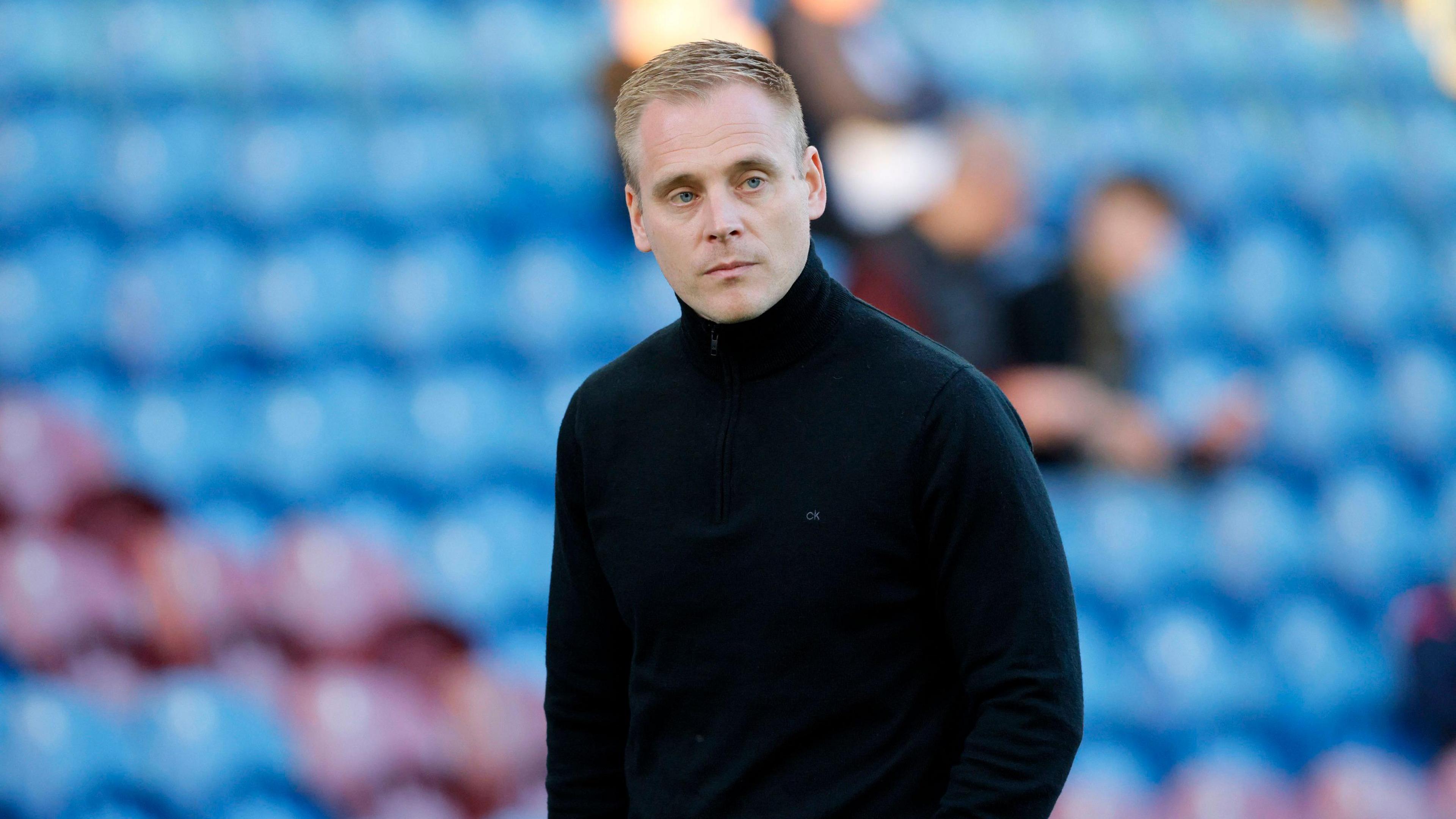 Johannes Hoff Thorup looking pensive and wearing black. He is standing in front of red and blue stadium seats.