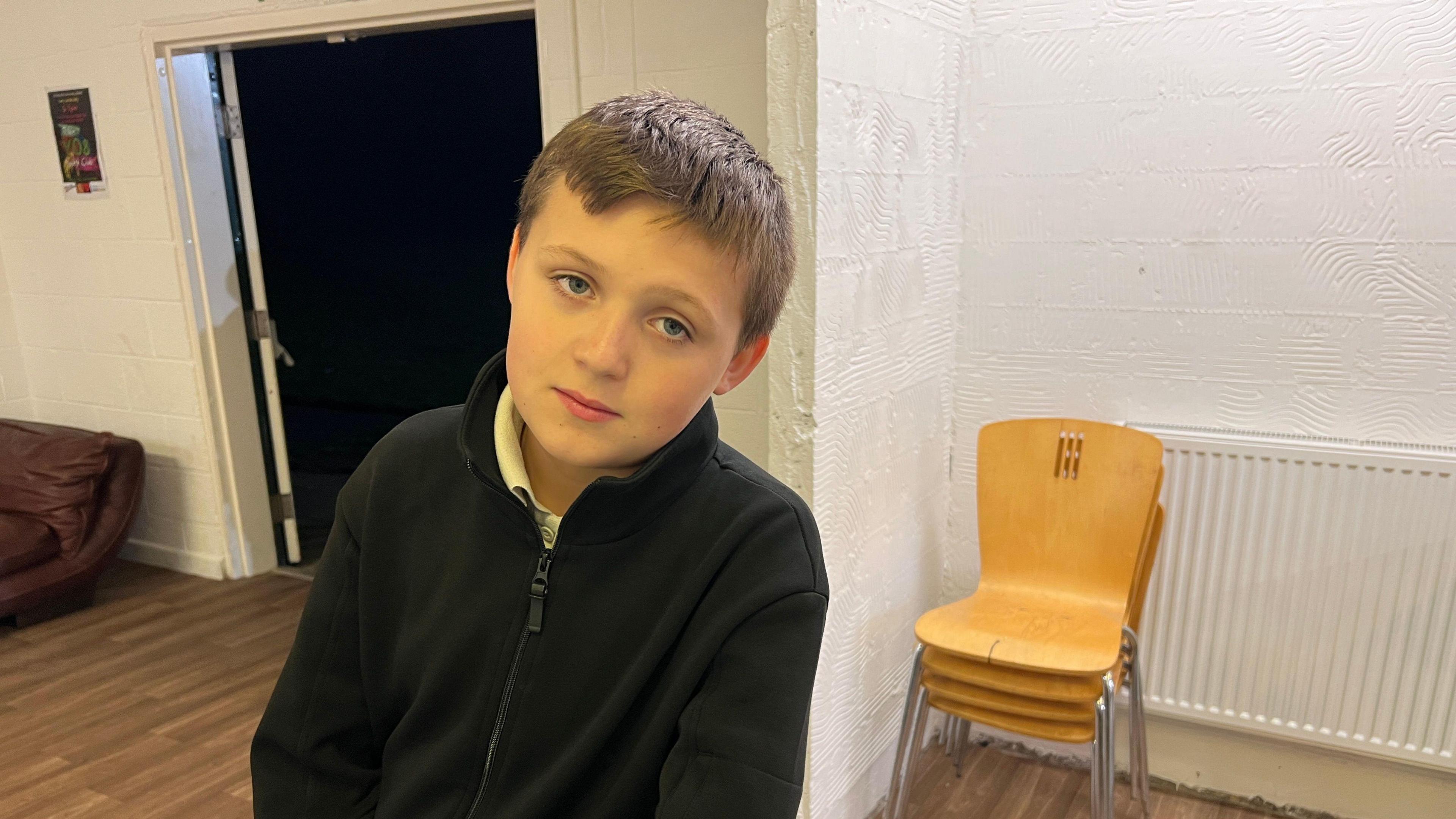 Caiden is leaning on a table and looking straight into the camera. He has brown hair and is wearing a black top. 