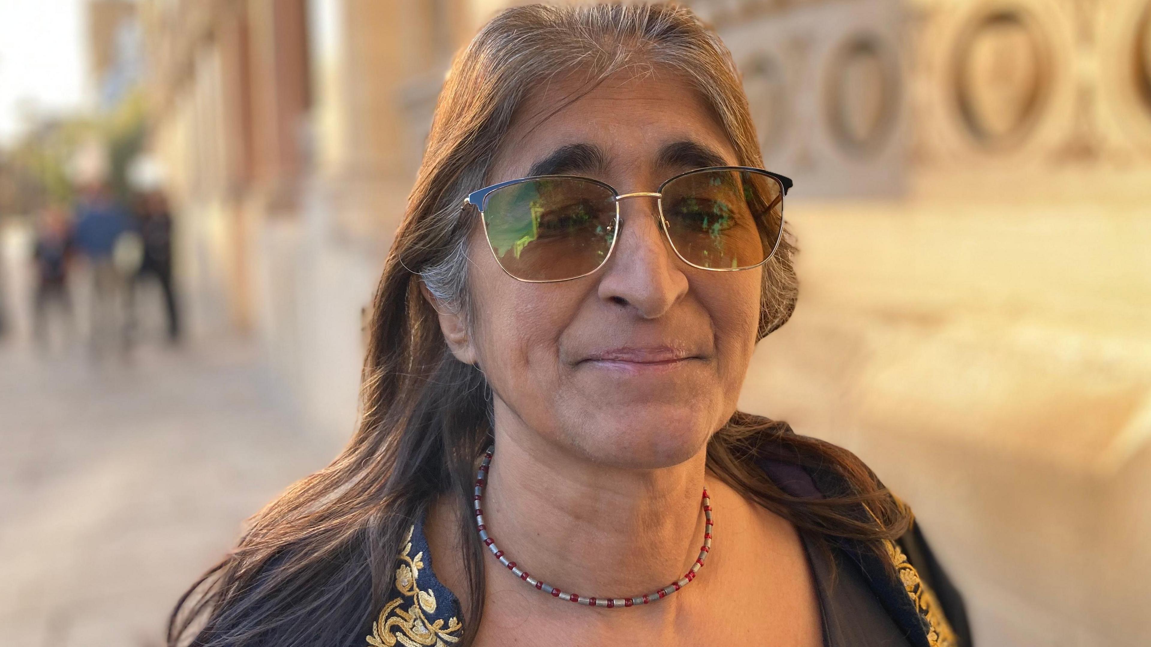 Anjona Roy, who has long dark hair and is wearing dark glasses and a black jacket with gold embroidery. She is standing outside Northampton's Guildhall. 