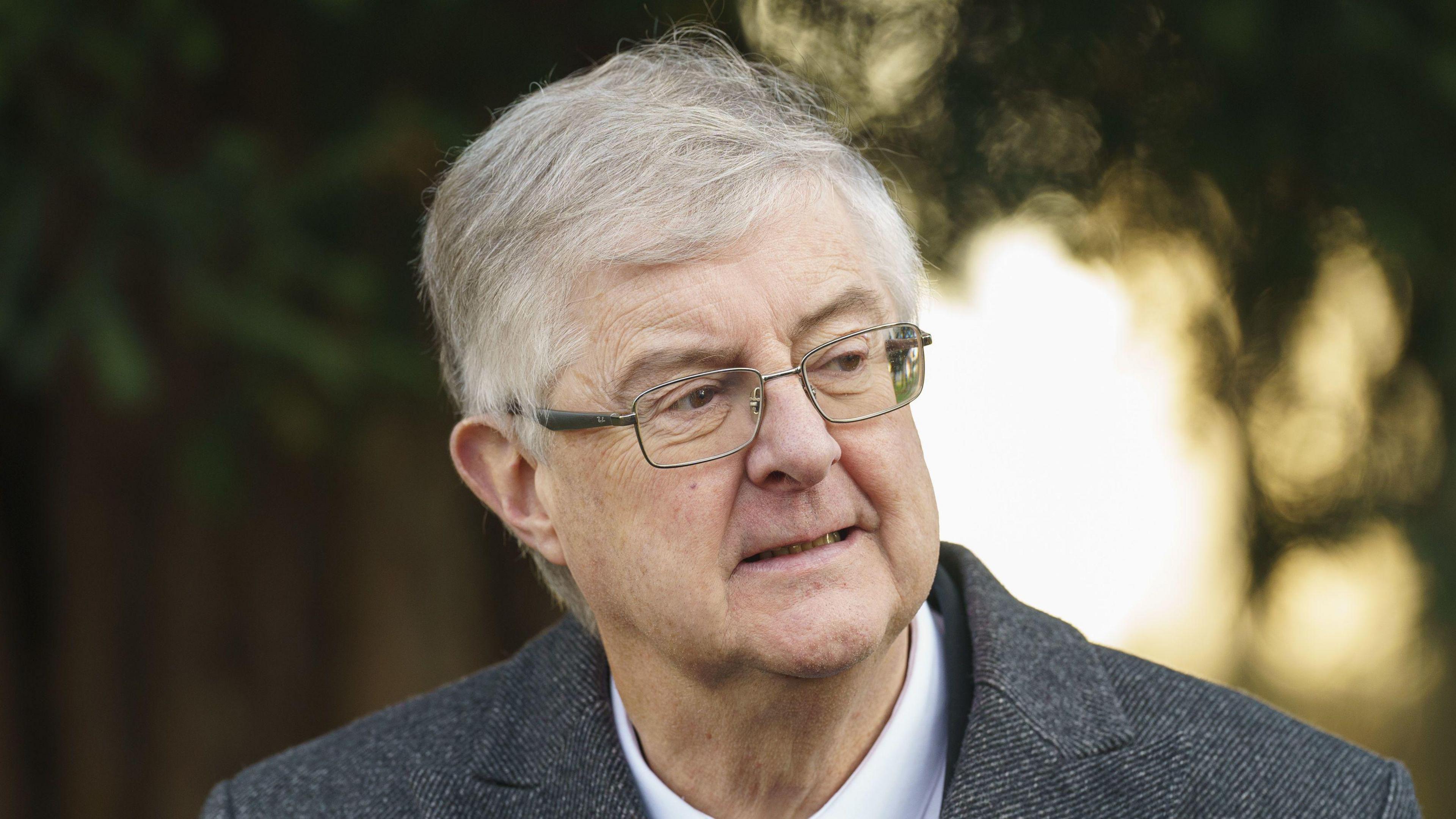 Mark Drakeford is wearing a grey suit, white shirt and glasses in front of a blurred background