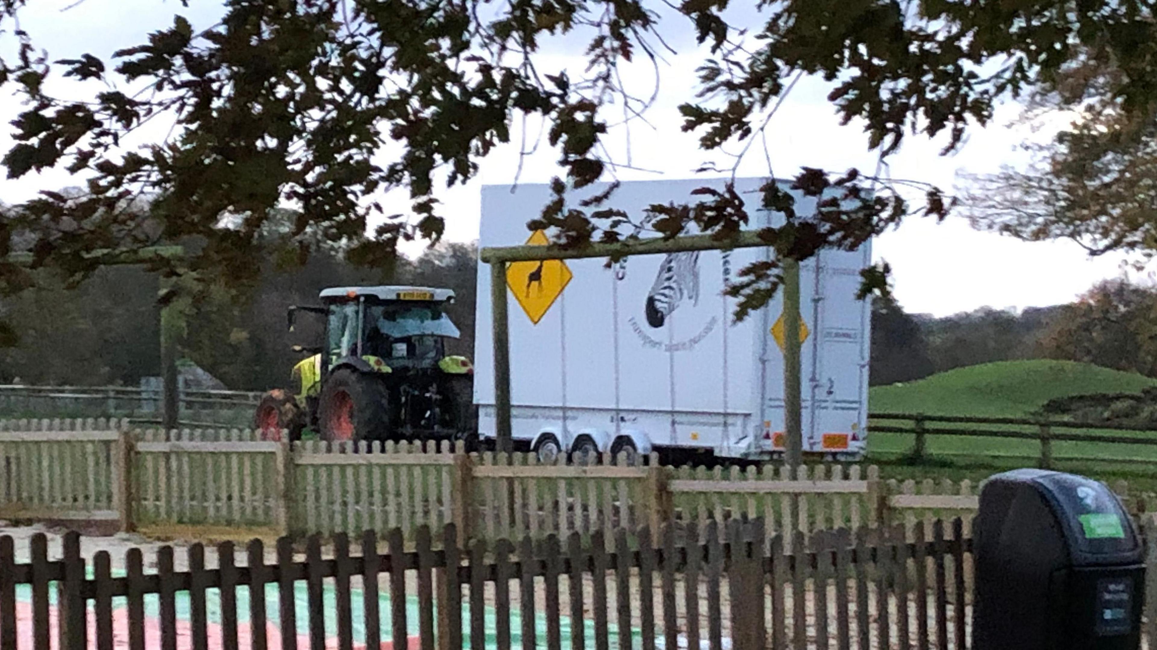 A green tractor is pictured with a large white trailor on the back of it. It is being moved through a field. The trailer has a giraffe logo printed on it as well as a zebra. 