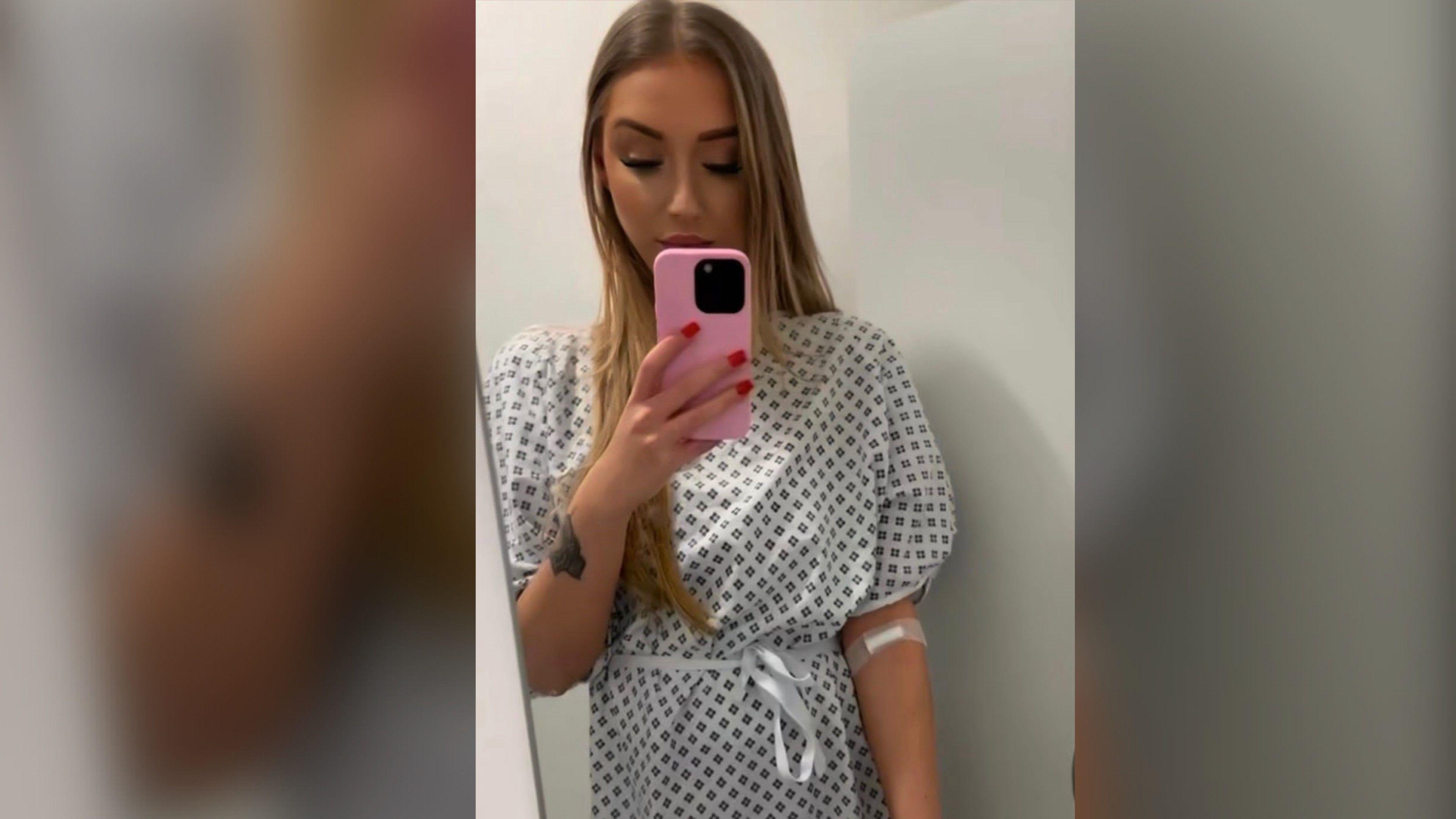 Ms Papadopoulos Green in a hospital gown, she has a plaster on her arm. She is taking a mirror selfie with her phone that has a pink phone case. 