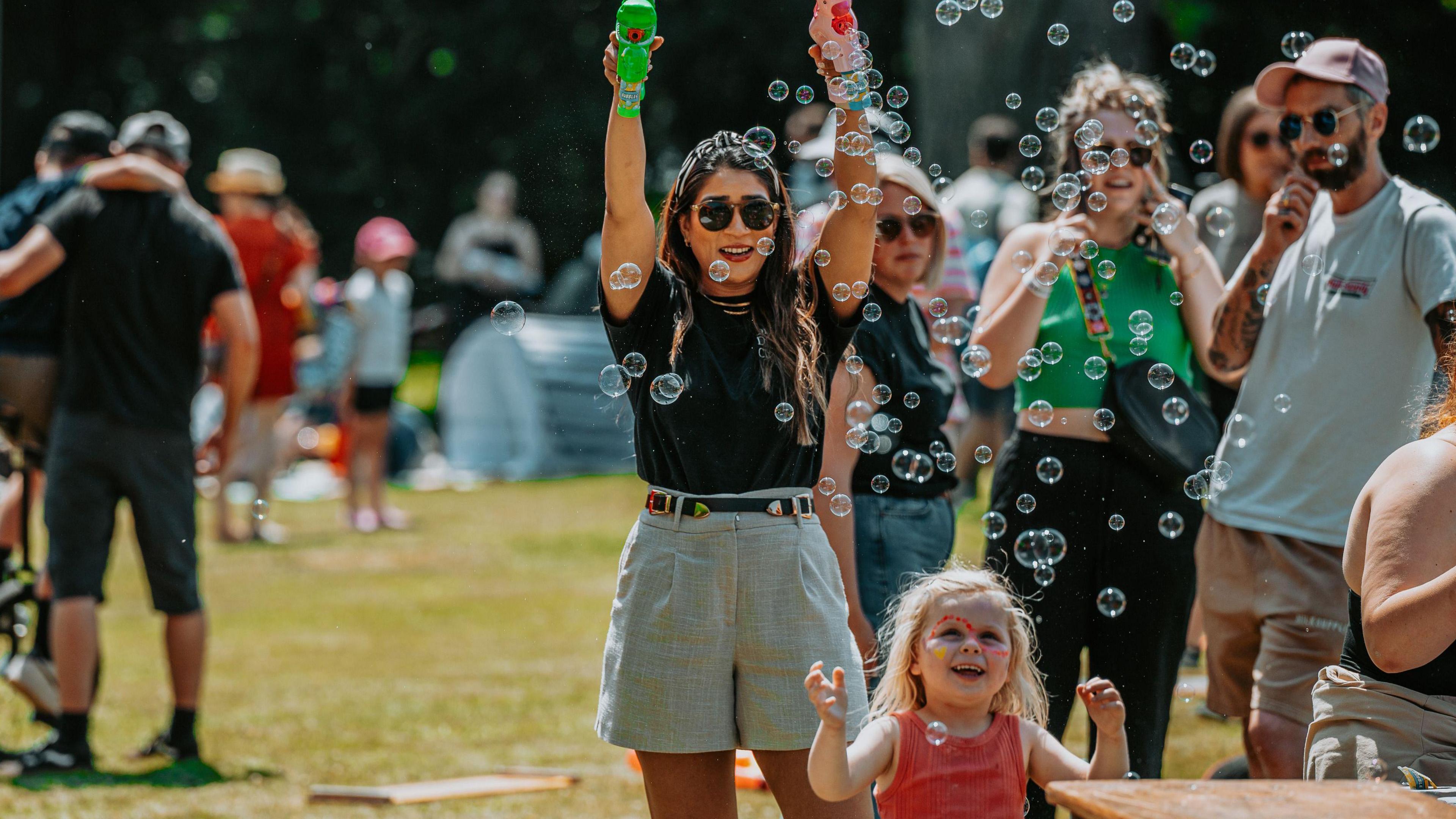 A woman in sunglasses holds up two bubble blasters and a small girl with facepaint on reaches up to the bubbles in the sunshine.