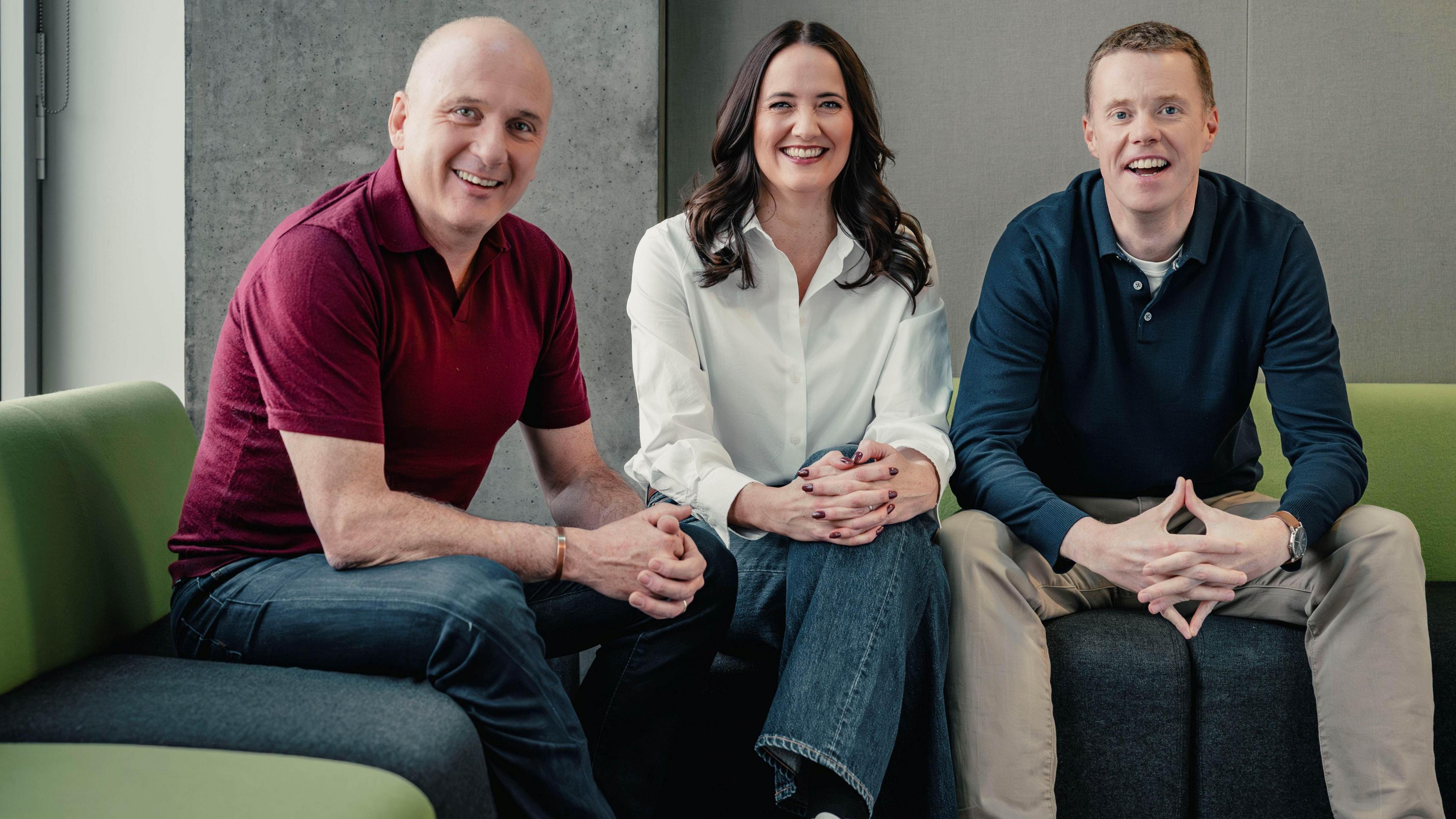 Martin Geissler, a bald man in a burgundy polo shirt and blue jeans, sits on a green sofa in a concrete-walled room. He is smiling. To his right Laura Maciver, a woman with long black hair, is wearing a white shirt and blue jeans and is also smiling. To her right sits Phil Goodlad. He is wearing a blue polo shirt and sand-coloured jeans and is also smiling.