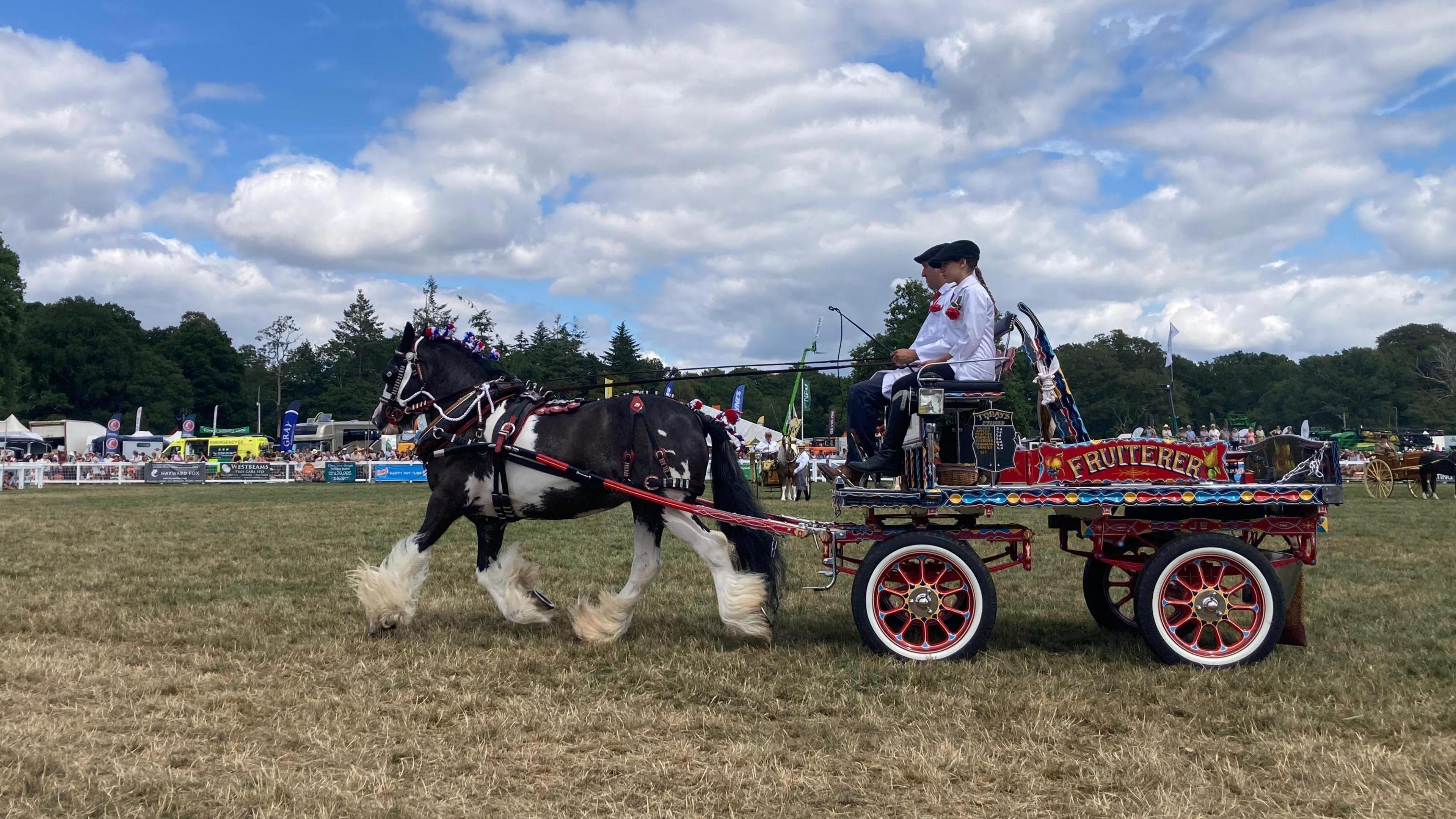 A horse and vintage fruiterers kart are driven around the New Forest Showground. Crowds and other stalls can be seen in the background.