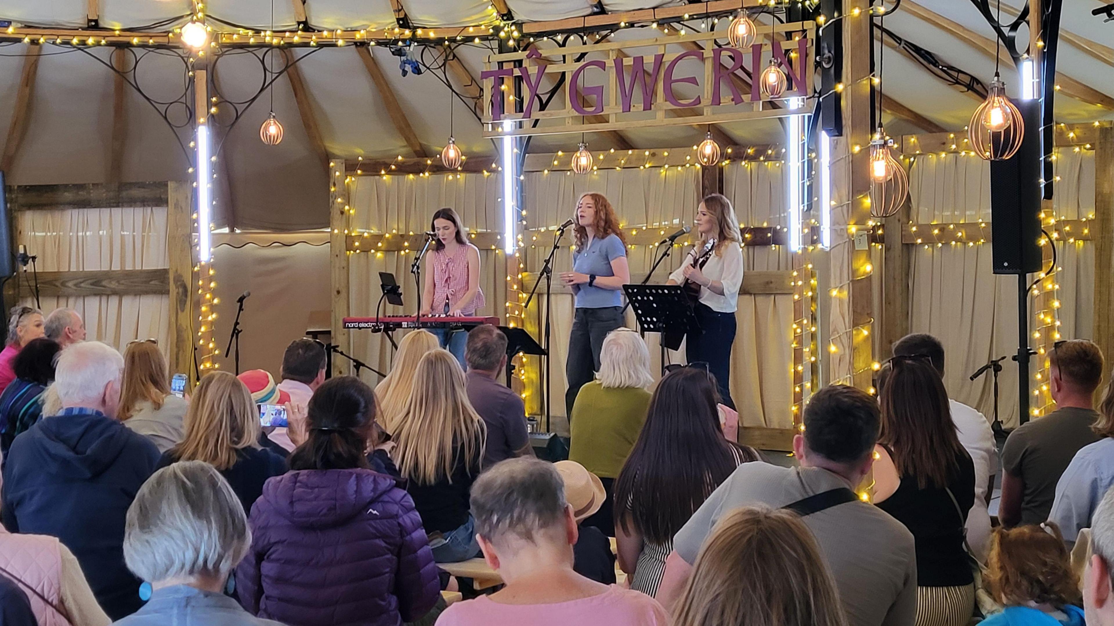 Singer flanked by one musician on guitar and another on keyboard perform beneath fairy lights before a seated audience