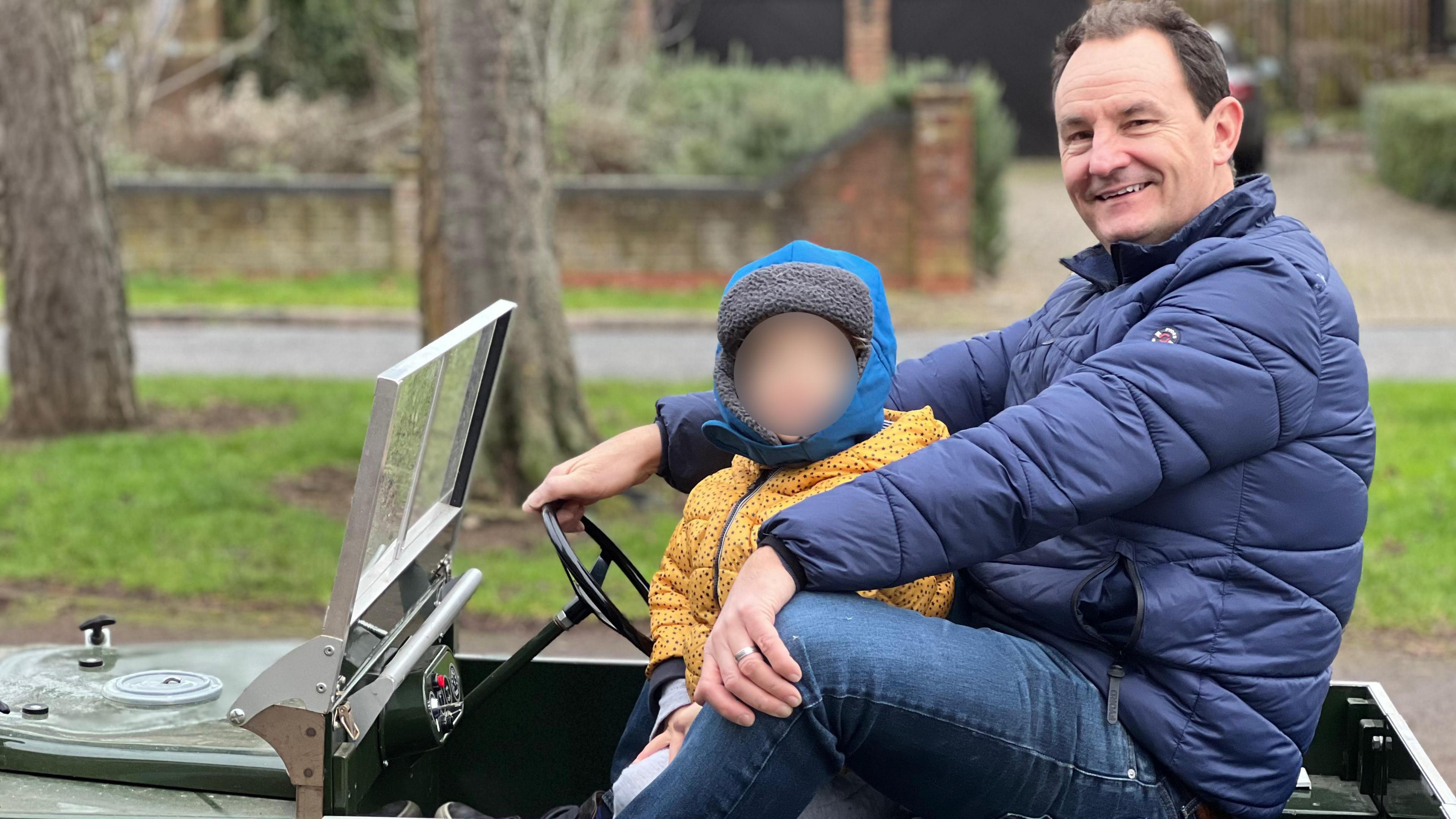 Philip Nash is sitting in the mini-Land Rover wearing a blue coat with his young son wearing yellow, sitting in front of him. The son's face has been blurred.