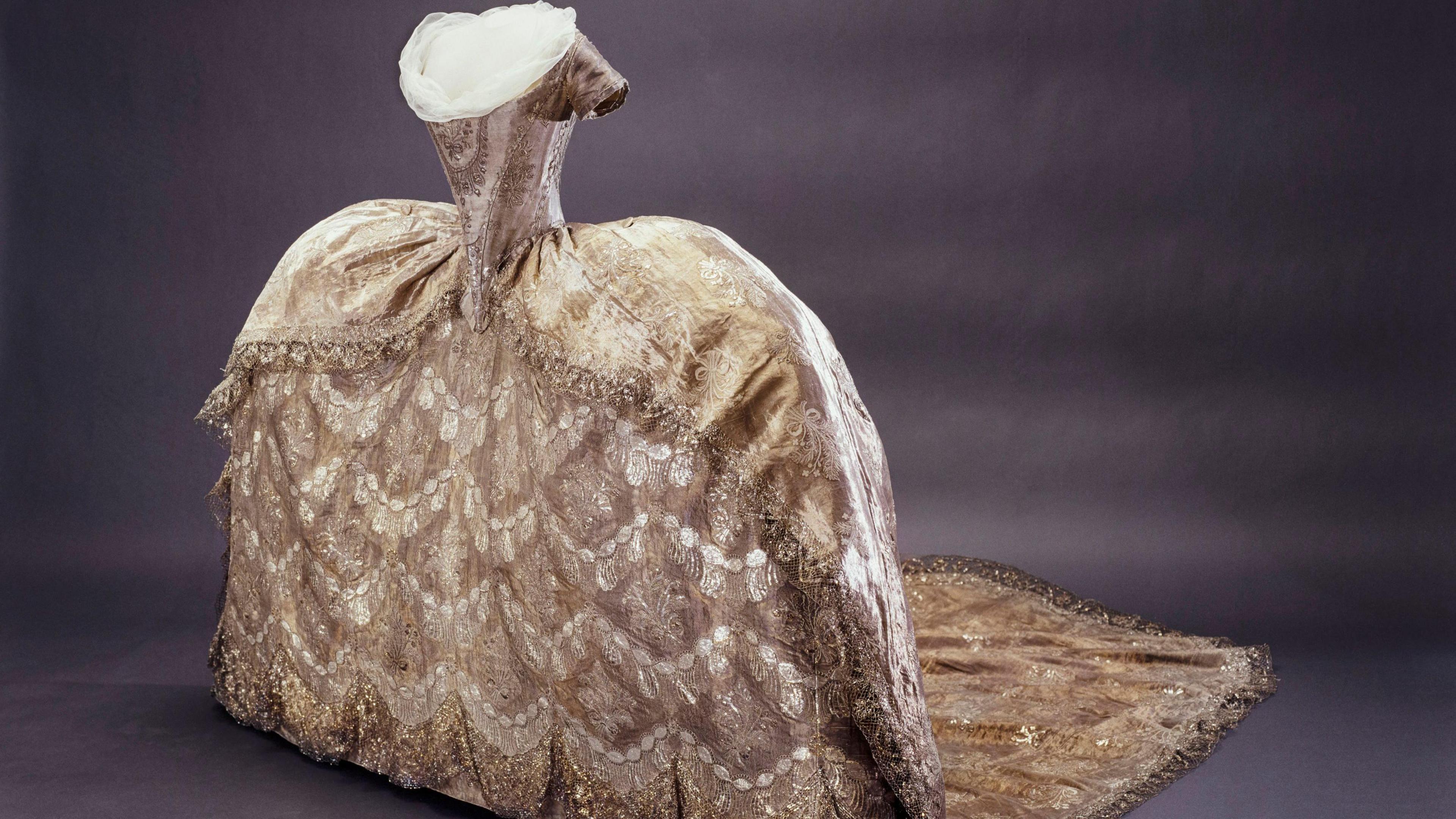 A luxurious court gown in gold and silver tones, featuring an extremely wide skirt supported by panniers, detailed with lace and metallic embroidery, displayed without a head or arms on a mannequin.