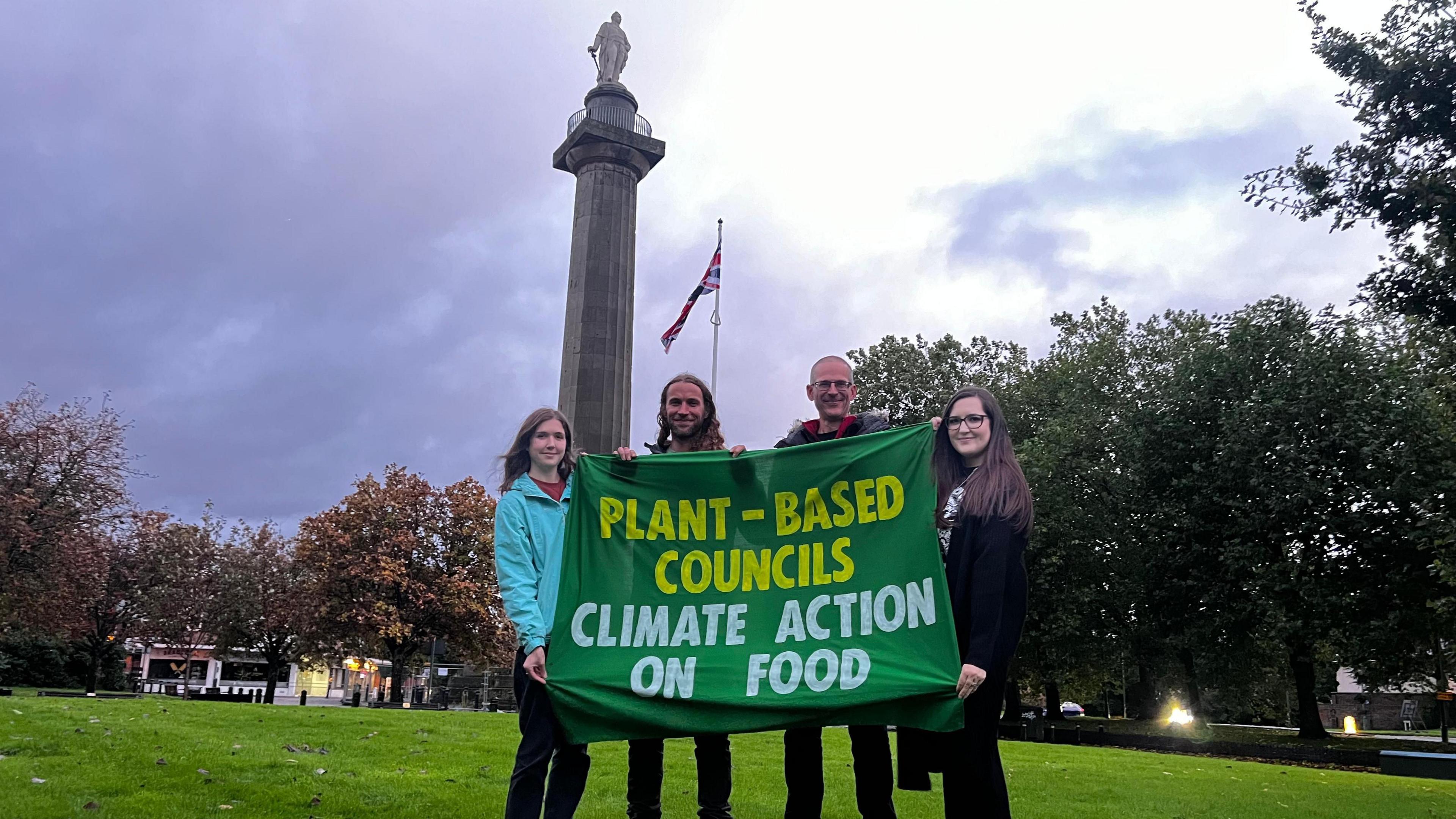 Four people are standing on grass in front of a large column with a statue of a man on top. They are holding a green banner that says "plant-based councils, climate action on food."