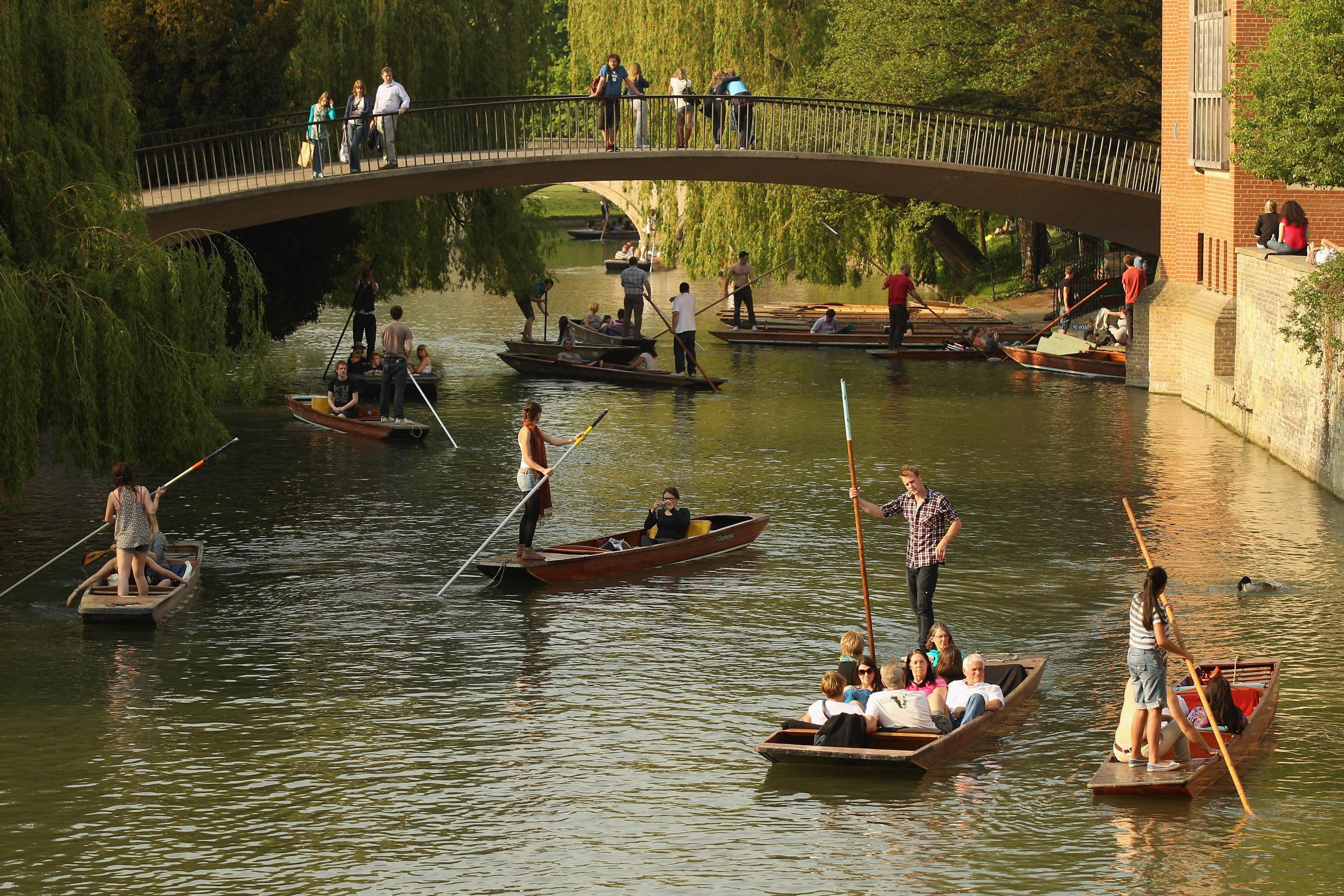 About seven punts on the River Cam in Cambridge. A footbridge with people on it crosses the river. Willow trees overhang the water.