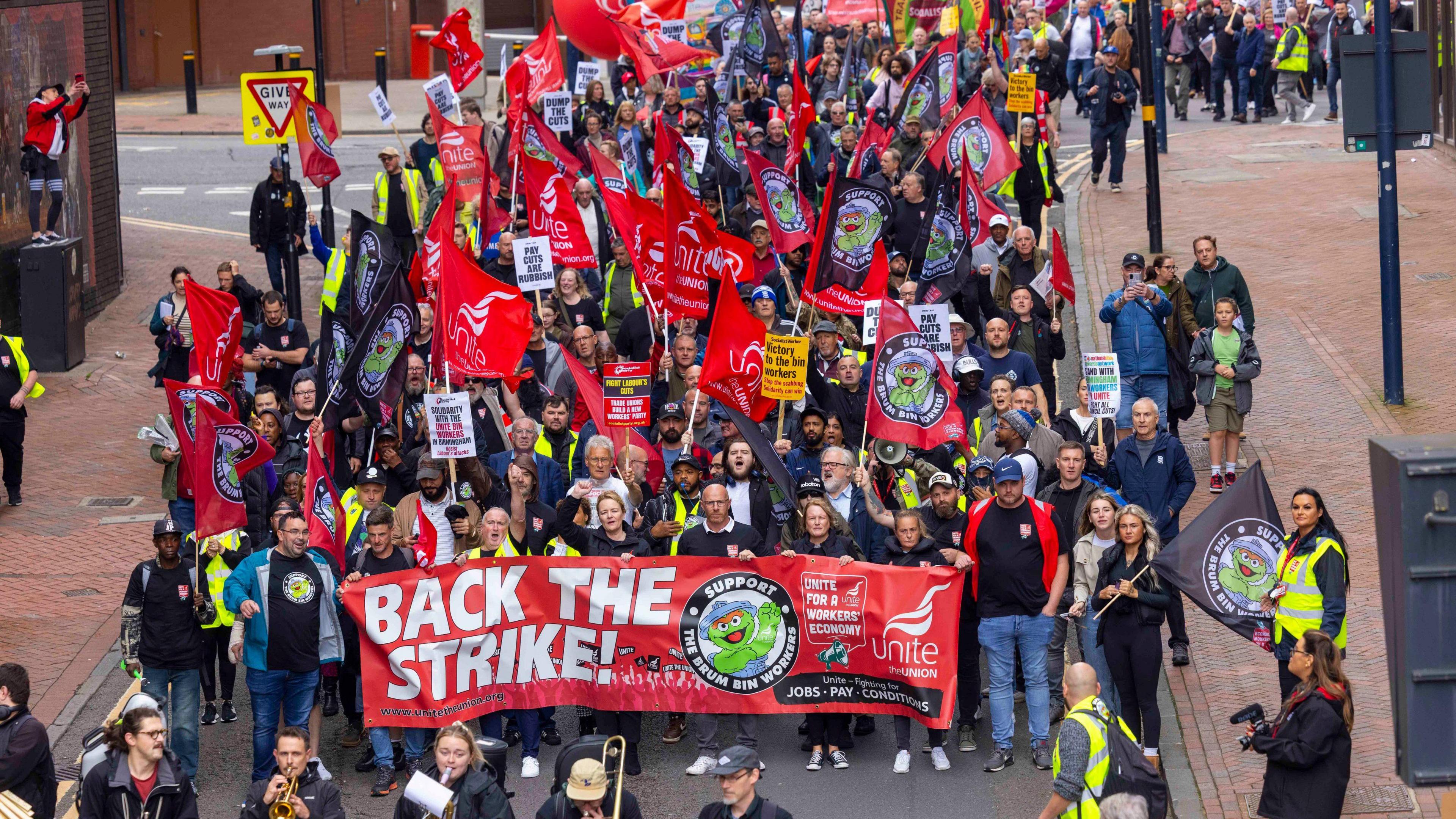 A crowd of people are holding red flags bearing the word Unite while the row at the front are holding a large banner bearing the words 'back the strike'