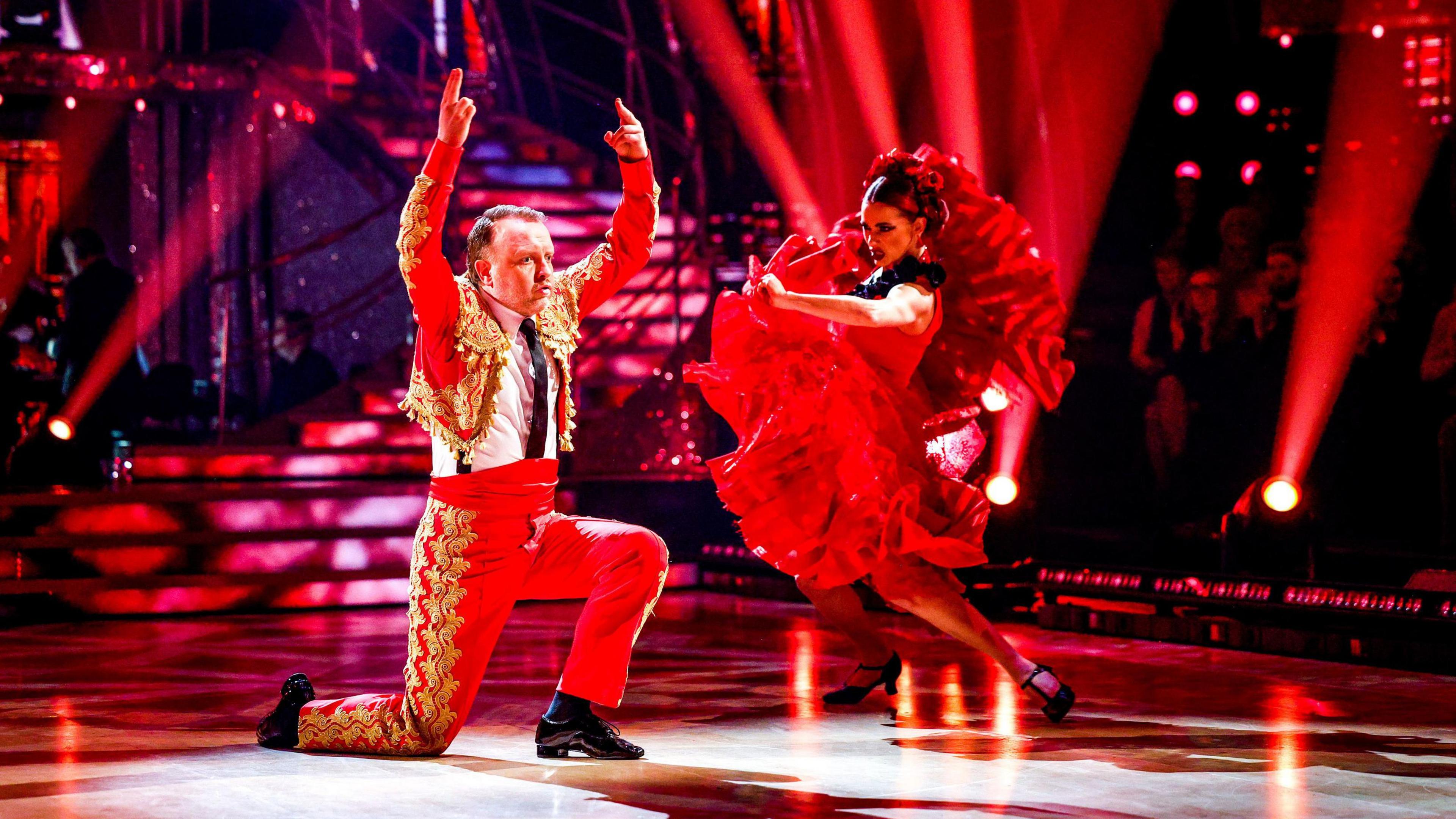 Chris McCausland and Dianne Buswell perform in red Flamenco-style outfits on the Strictly Come Dancing dance floor. He has slicked-back brown hair, and is down on one knee with his arms in the air. She has her hair tied back with flowers in, and is holding up the bottom of her dress as she dances next to him. 