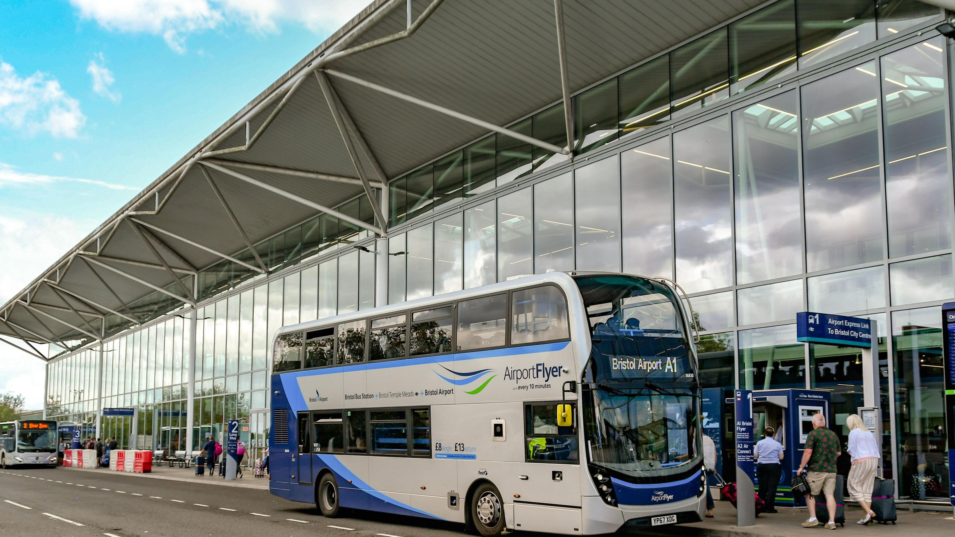 People queueing for bus outside Bristol Airport, which has big glass windows and other buses in the distance.
