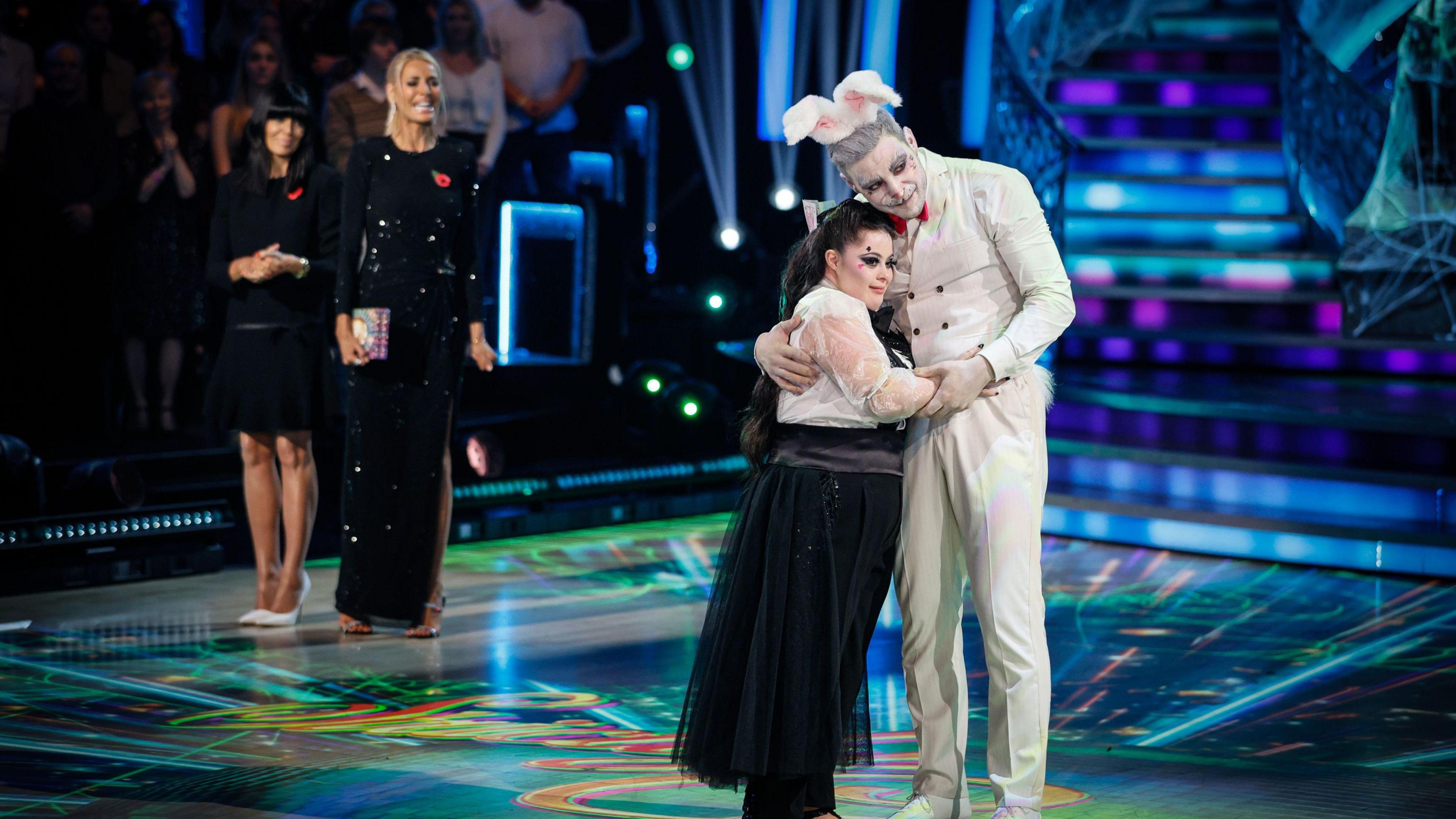Ellie Goldstein, who is dressed in a long black dress and a white shirt, is hugging her partner Vito who is dressed as a bunny rabbit in a white suit. They are standing on the dancefloor of Strictly Come Dancing.