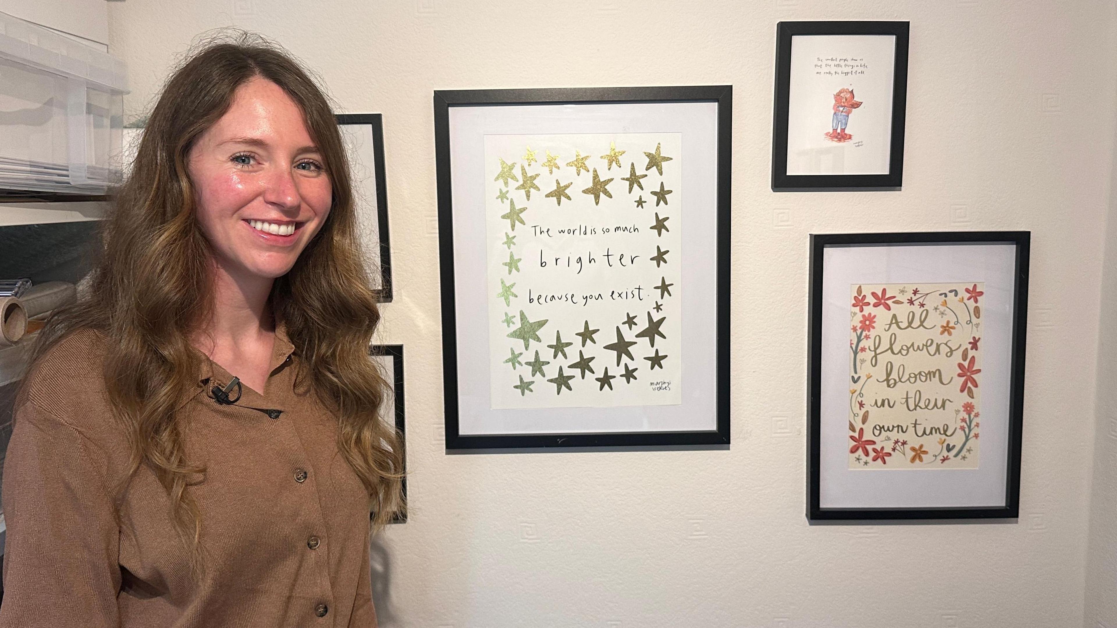 Kerri Cunningham is smiling at the camera in her office. She is standing in front of a cream wall where five of her sketches are hanging in black photo frames. One reads: 'All flowers bloom in their own time' while another reads: 'The world is so much brighter because you exist'.