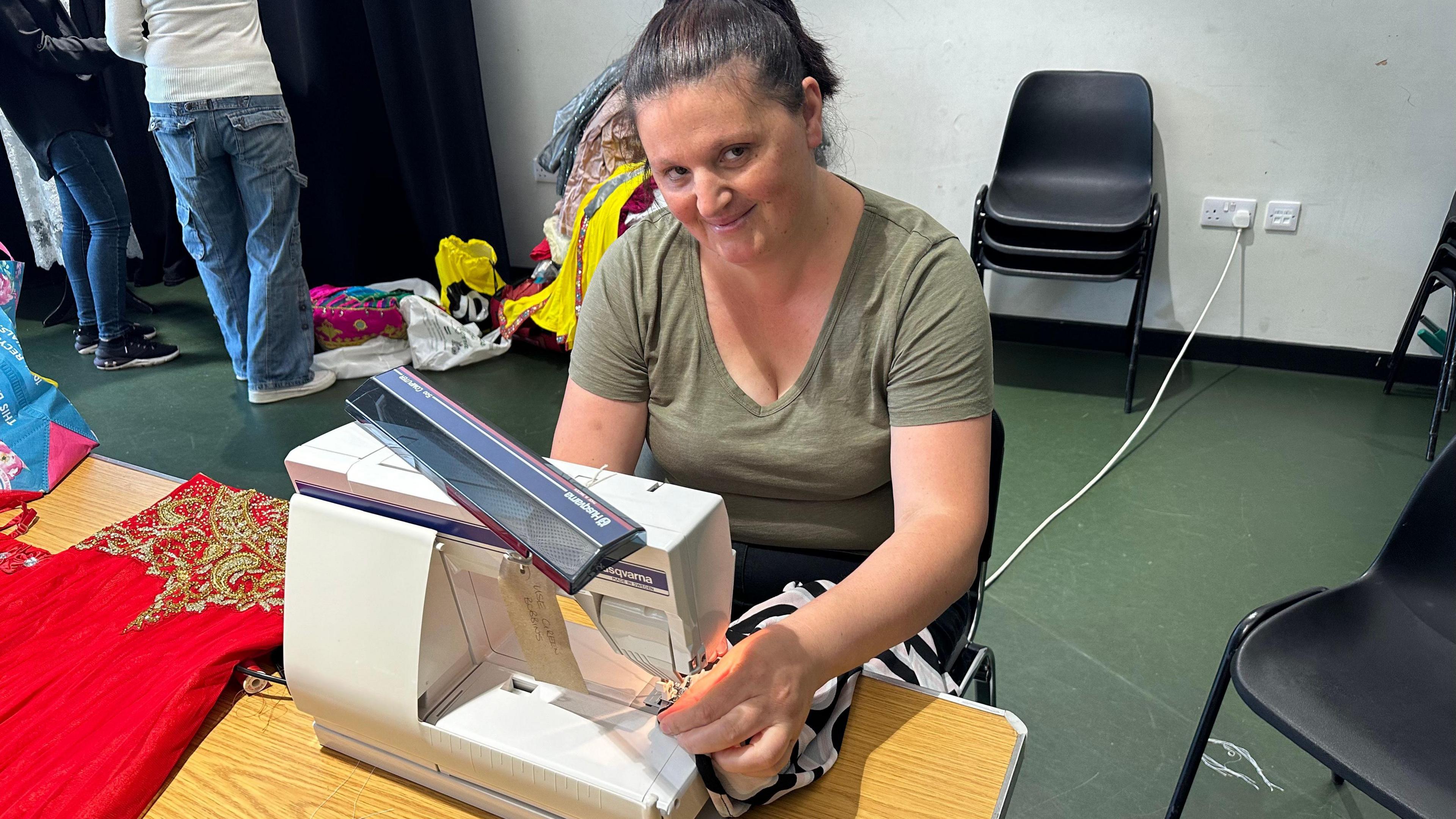 Fatimire Ibrohimi, wearing a khaki v-neck top smiling at the camera. She is sad behind a sewing machine and is putting a black and white stripy garment up to the machine's needle