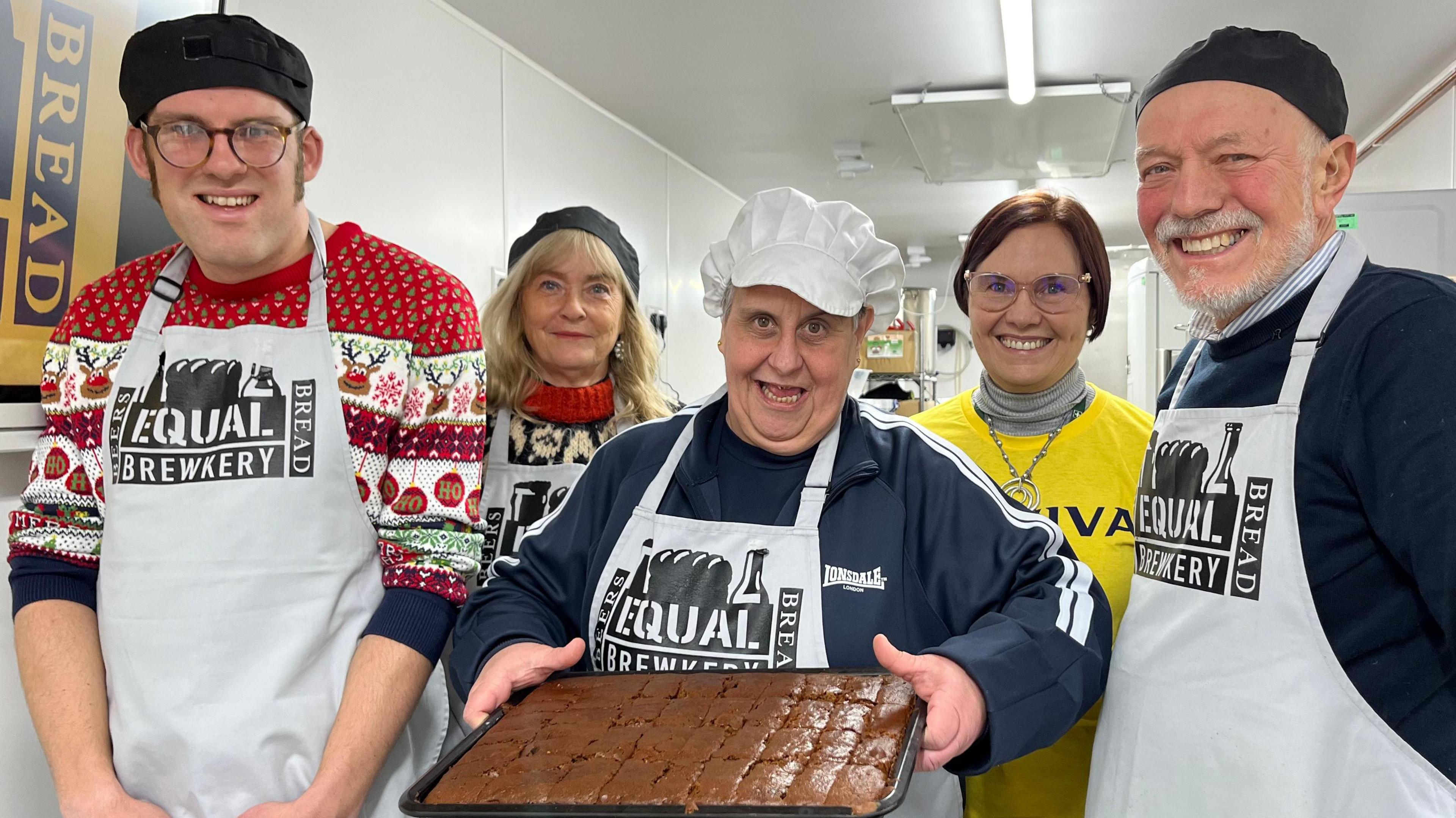 Five people stand inside a kitchen. Four are wearing aprons and bakers' hats. The woman in the middle is holding a tray of freshly-baked items, possibly gingerbread. All are smiling.