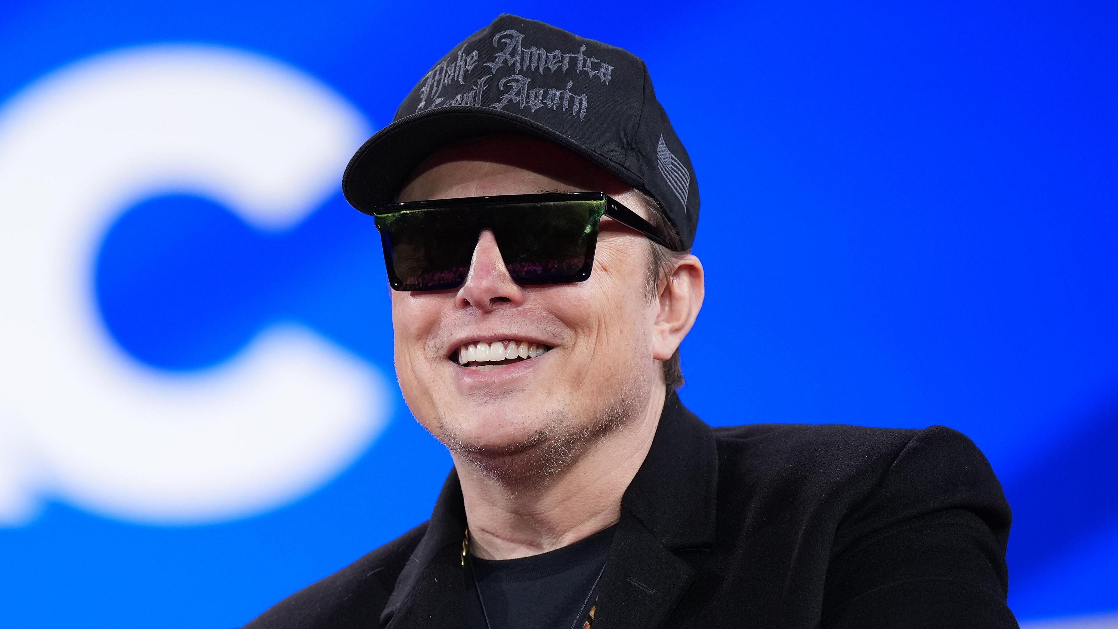 Tech multi-billionaire Elon Musk speaking at the Conservative Political Action Conference (CPAC) in February 2025. He is dressed all in black, including a Make America Great Again baseball cap, coat, t-shirt and sunglasses. The background is bright blue with a large white letter C over Musk's right shoulder.