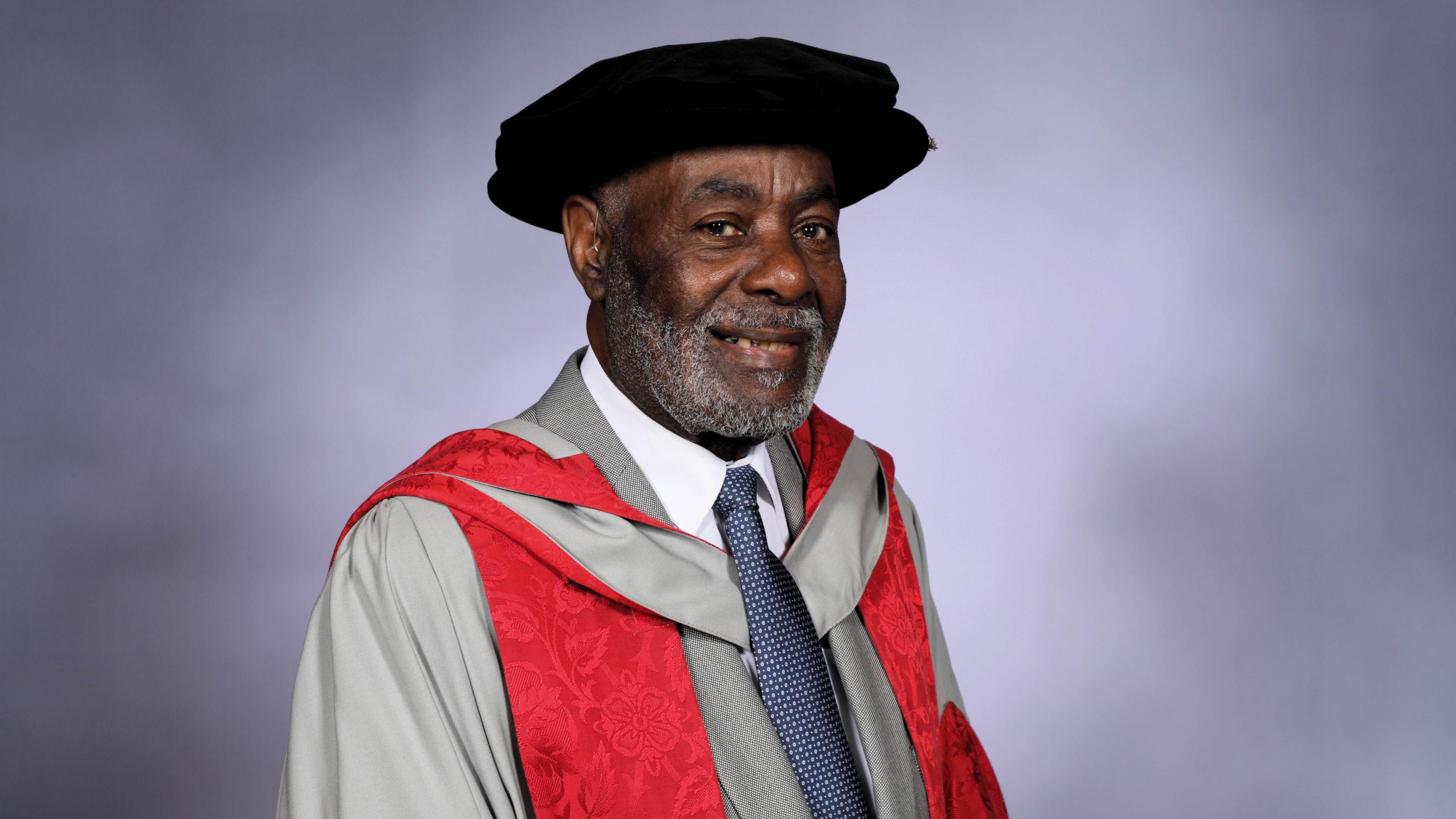 Guy Bailey OBE. He is pictured wearing a graduation cap and gown. He is smiling at the camera. 