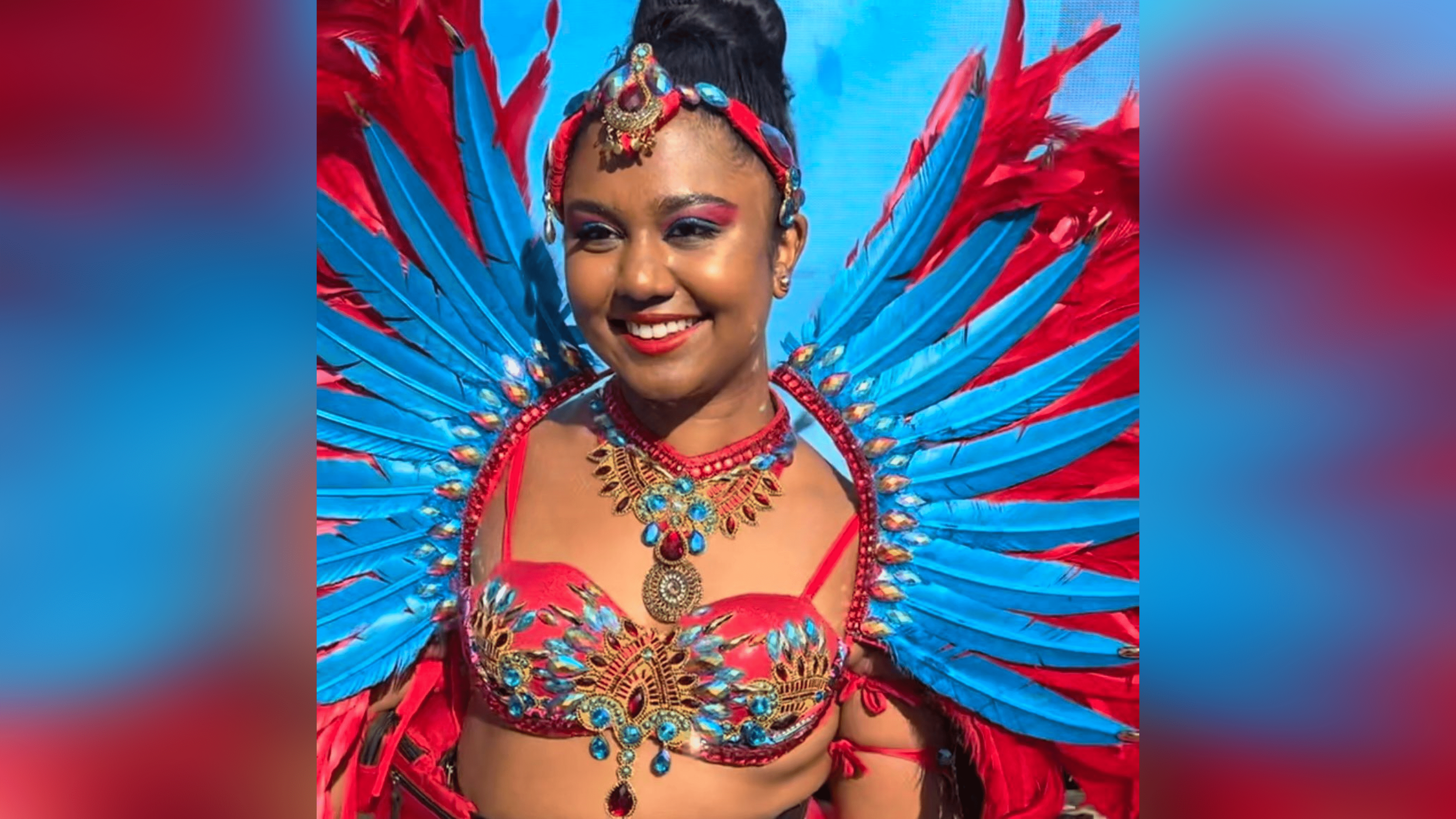 Sophia Estelle Mangroo in a red and blue carnival outfit. It includes a vibrant spread of feathers behind her back, and an intricately beaded bra-style top, with a jewelled necklace and headpiece to match. She also has red and blue eyeshadow on, and red lipstick. Her hair is in a bun on top of her head.