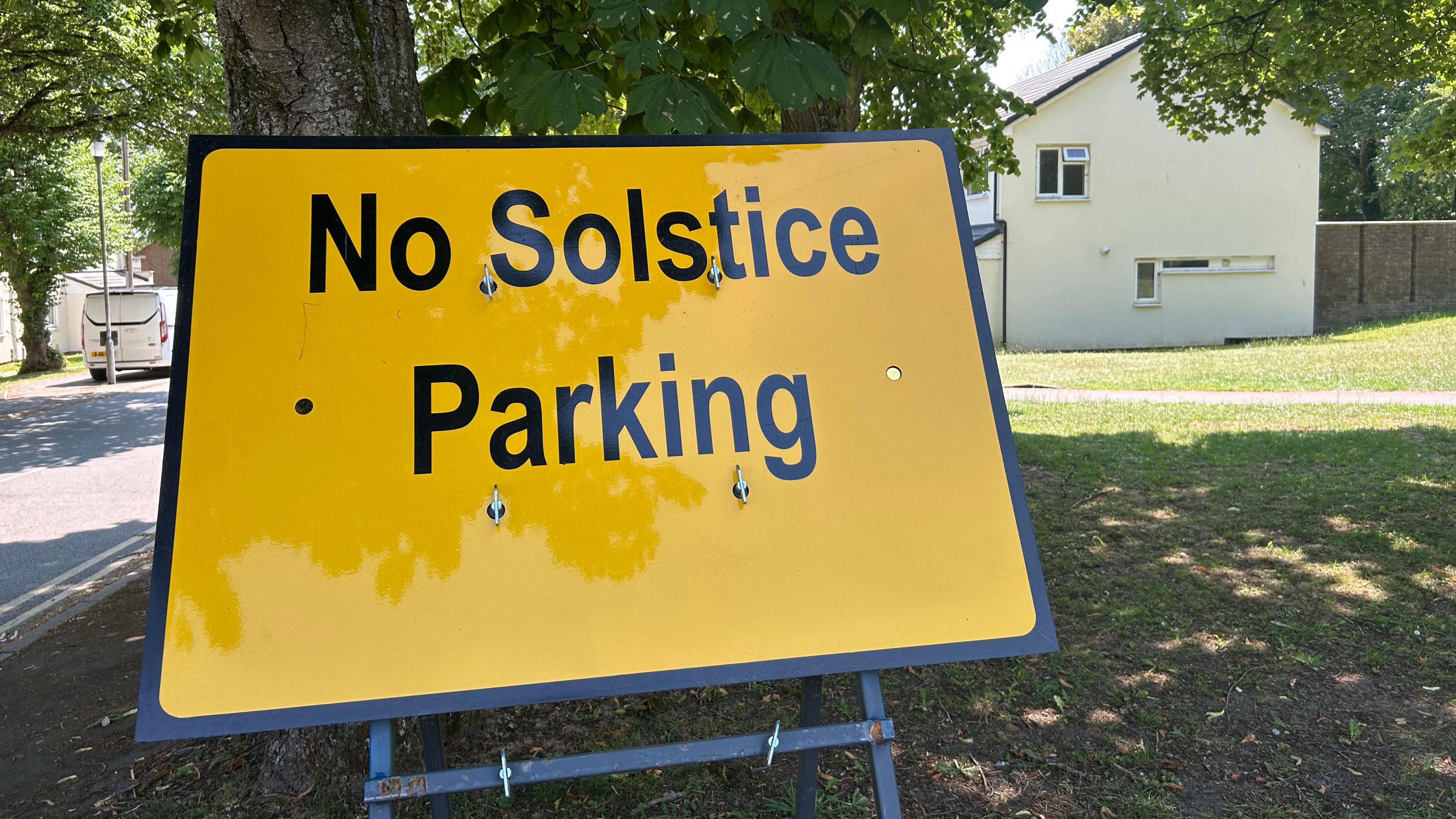 A large yellow temporary sign reading "no solstice parking" is on a piece of grass underneath a tree. A couple of homes can be seen.