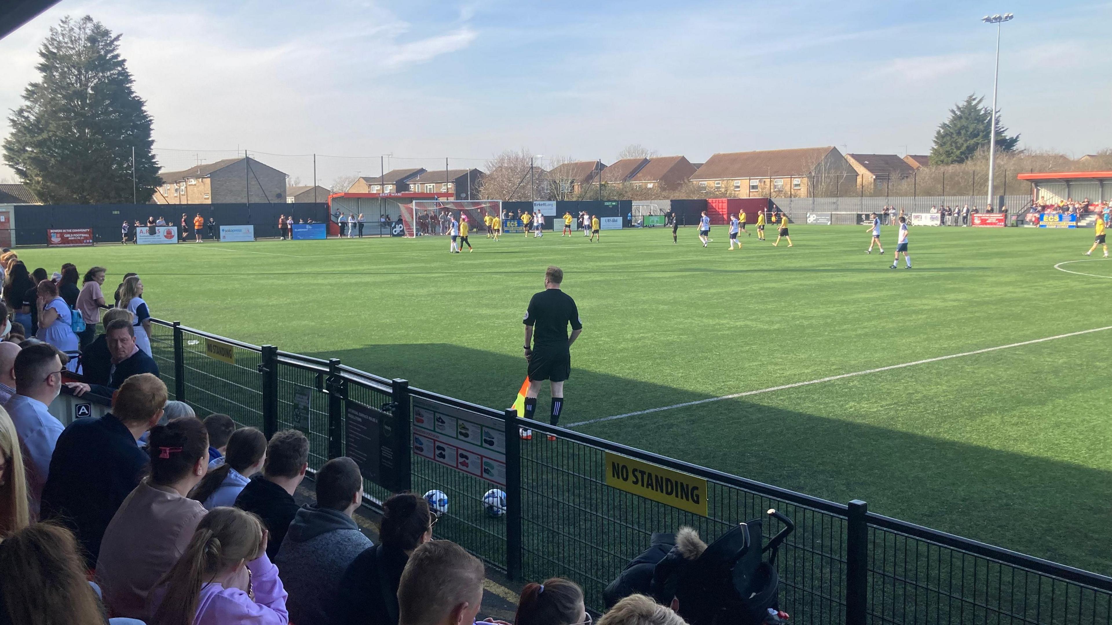 Fans watching a football match. Spectators are sitting on the left behind a low back fence. An assistant referee is standing in the centre wearing a black top and shorts and holding an orange and yellow flag. One the right players wearing  yellow tops and white and blue tops on taking part in a match on a football field.