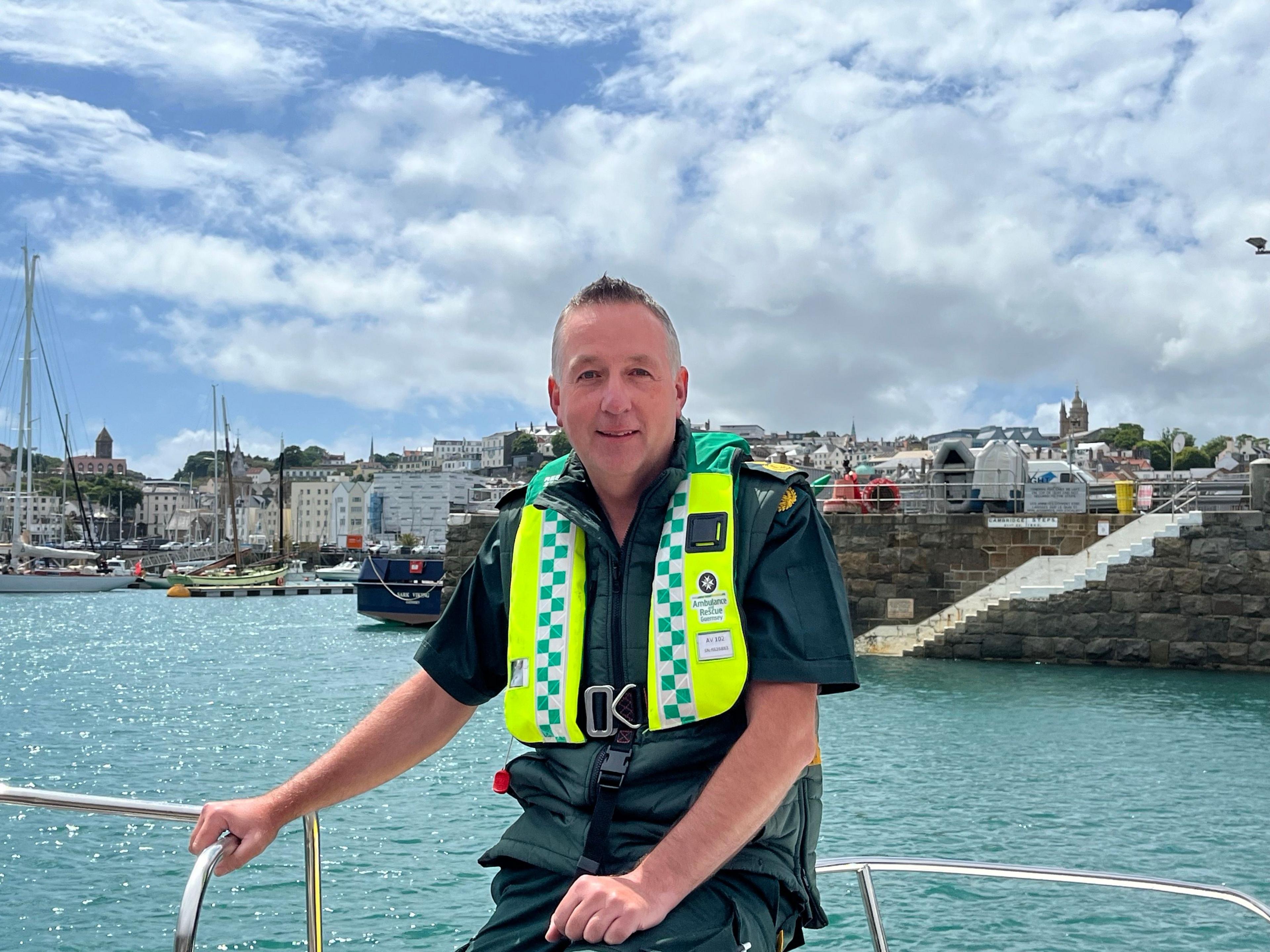 A man in a green ambulance uniform with a high-visibility life jacket sits on a boat in a rural harbour.