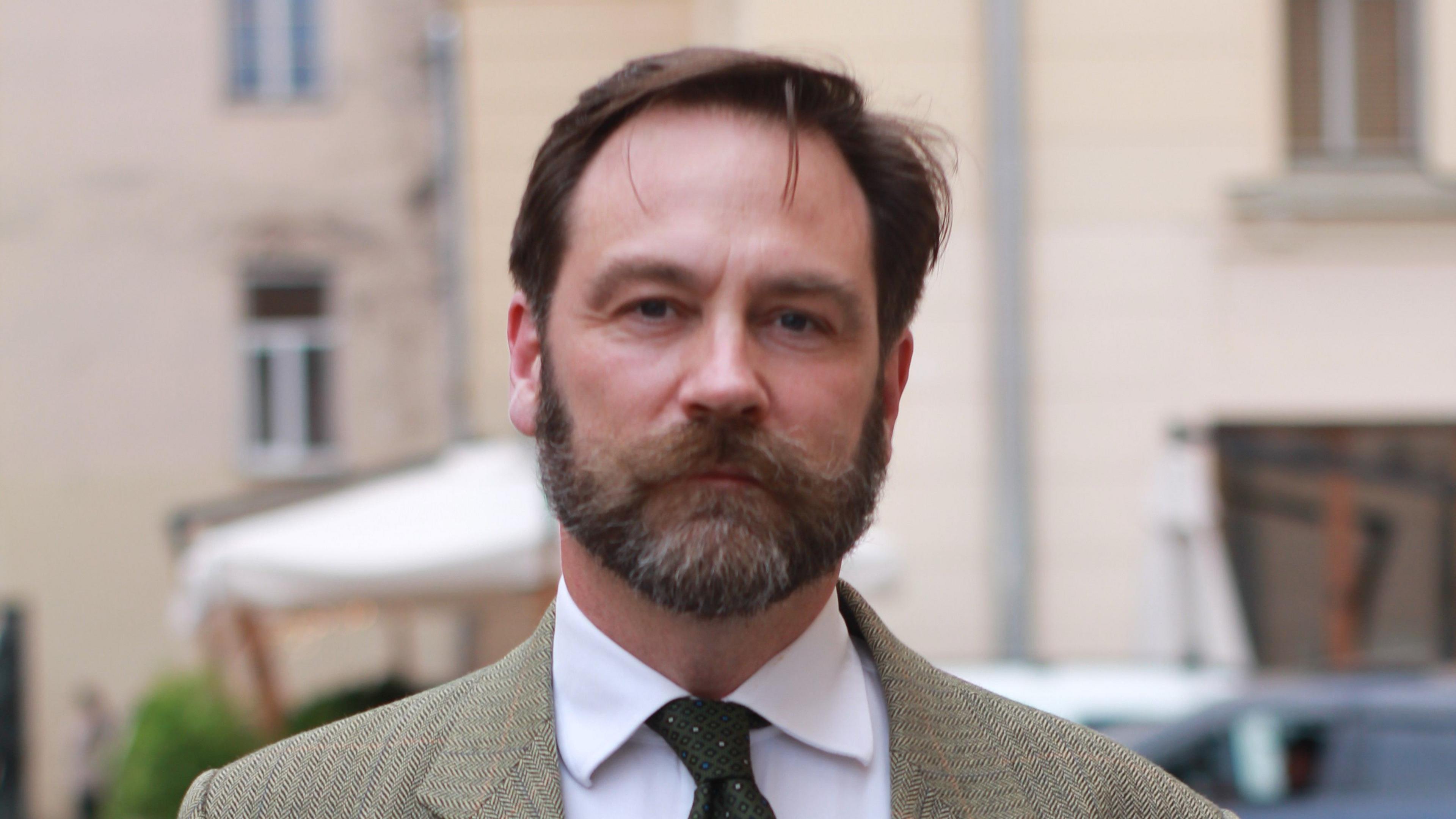 A man looking at the camera. He has dark hair and a thick dark beard.