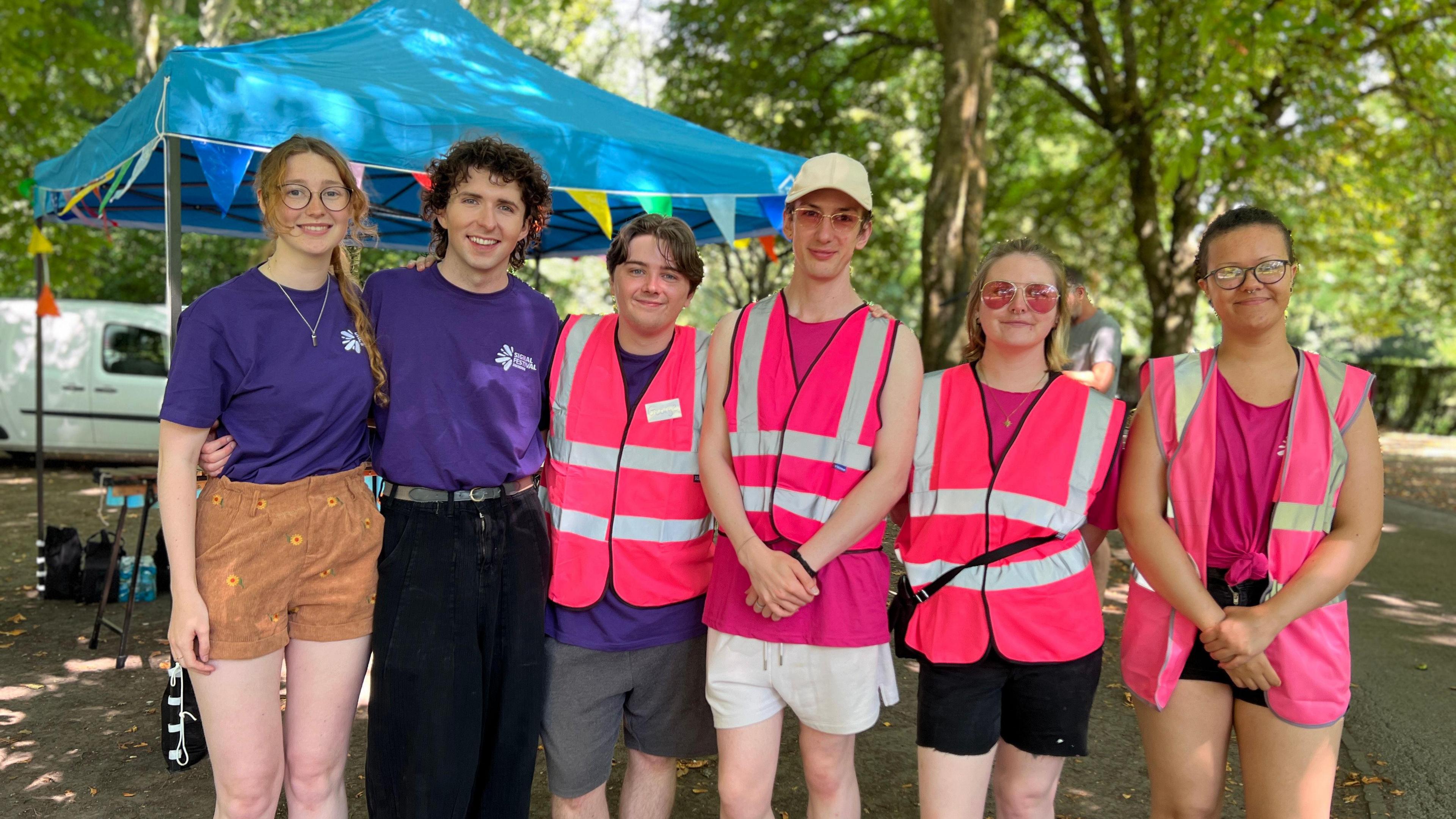 Six people, four in high-vis pink jackets and two in purple t-shirts, smiling at the camera