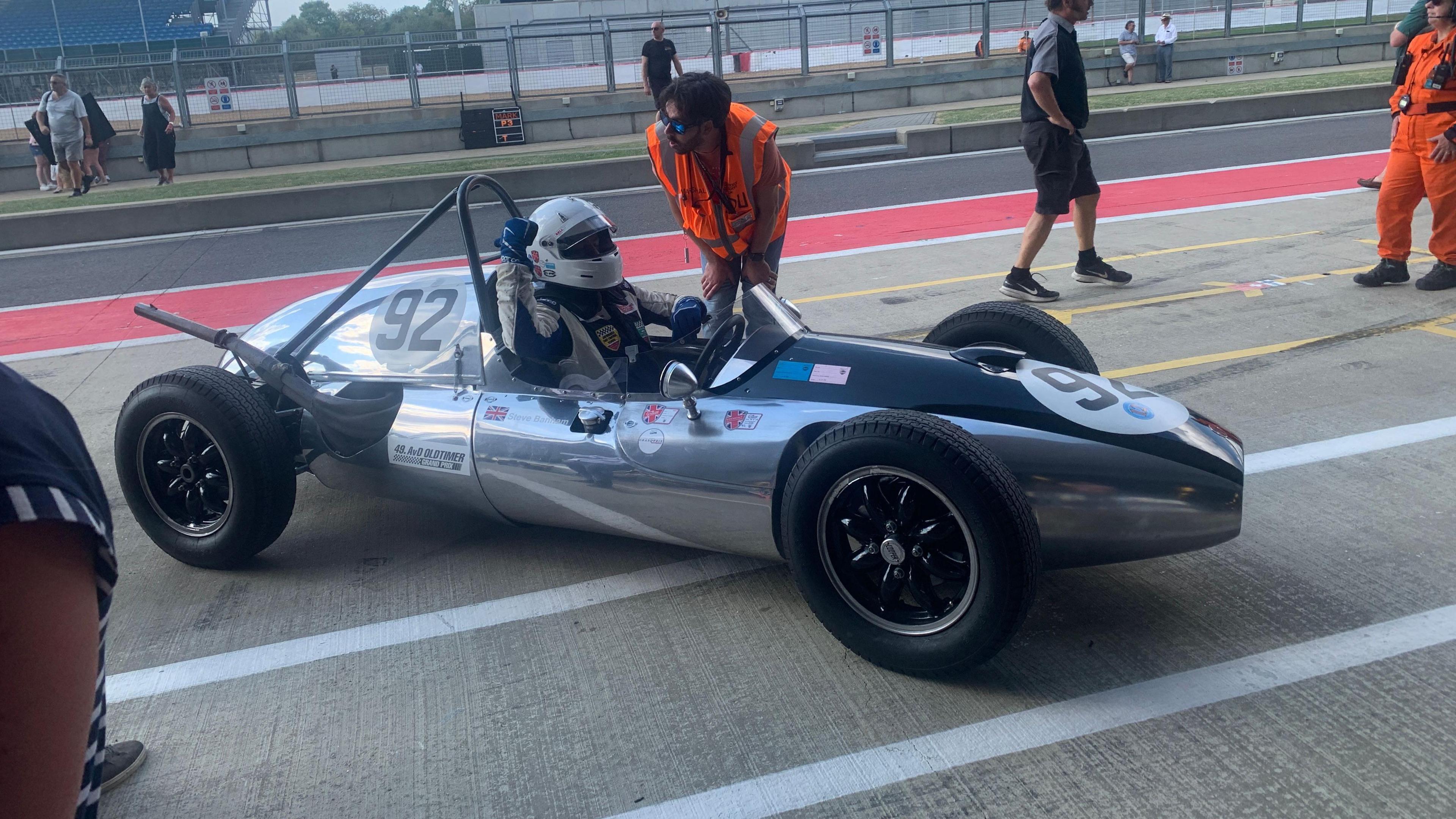 A racing car with someone sat inside it. There are people around the car.