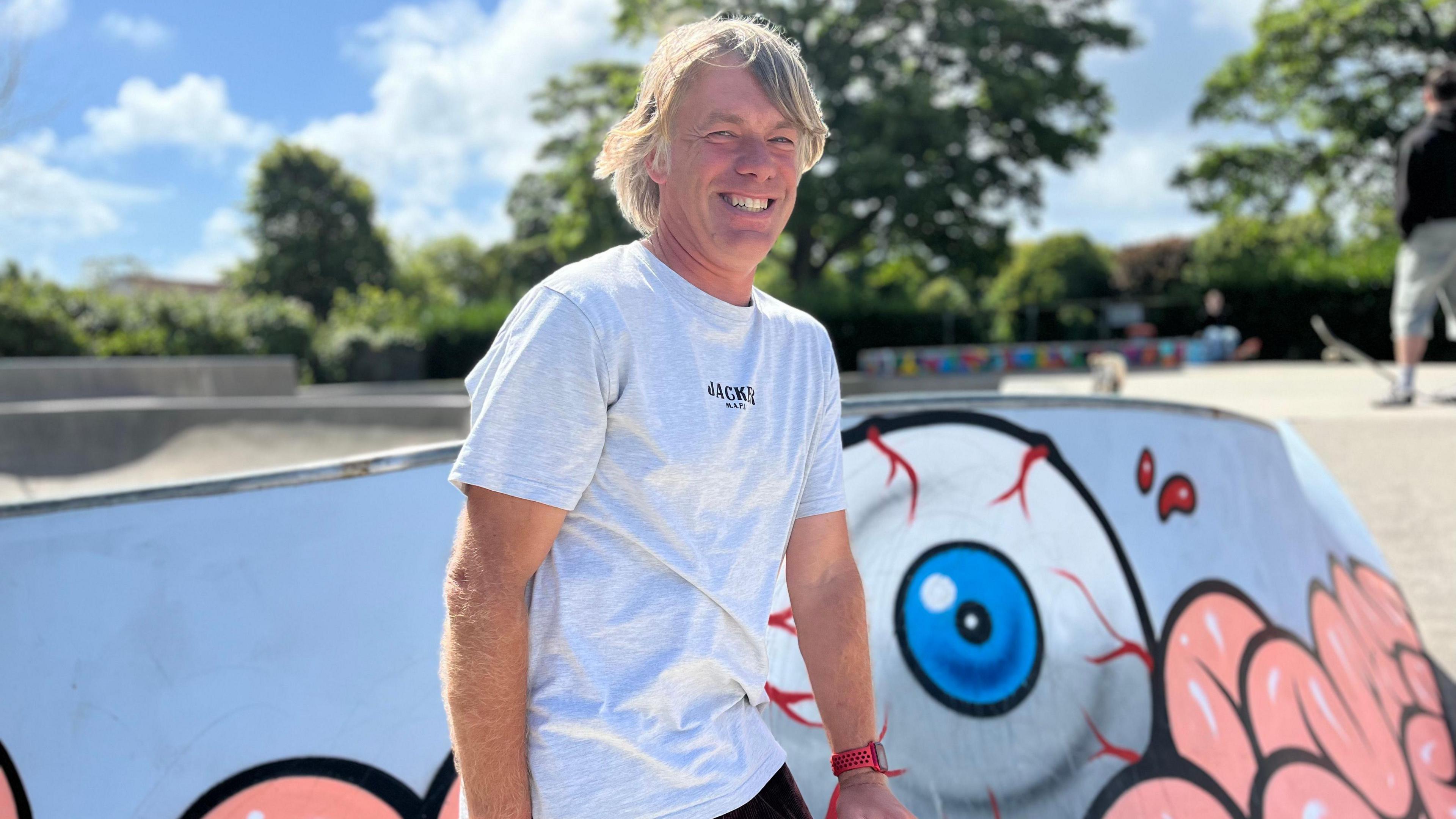 A smiling man in a t-shirt holds a skateboard in a skatepark.
